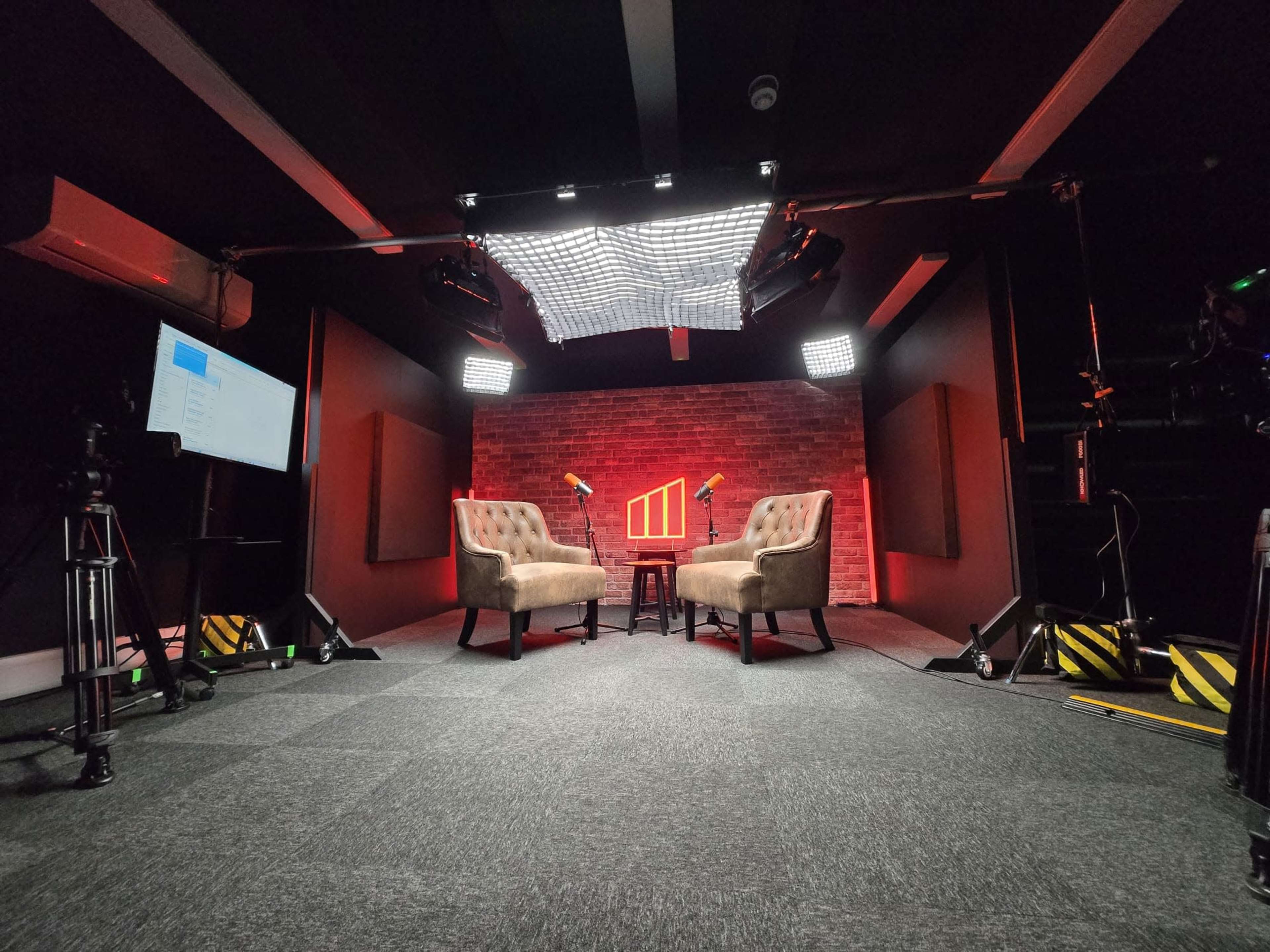 The image shows a dimly lit studio setup with two armchairs facing each other, a small table between them, and multiple lights and cameras positioned around the space.