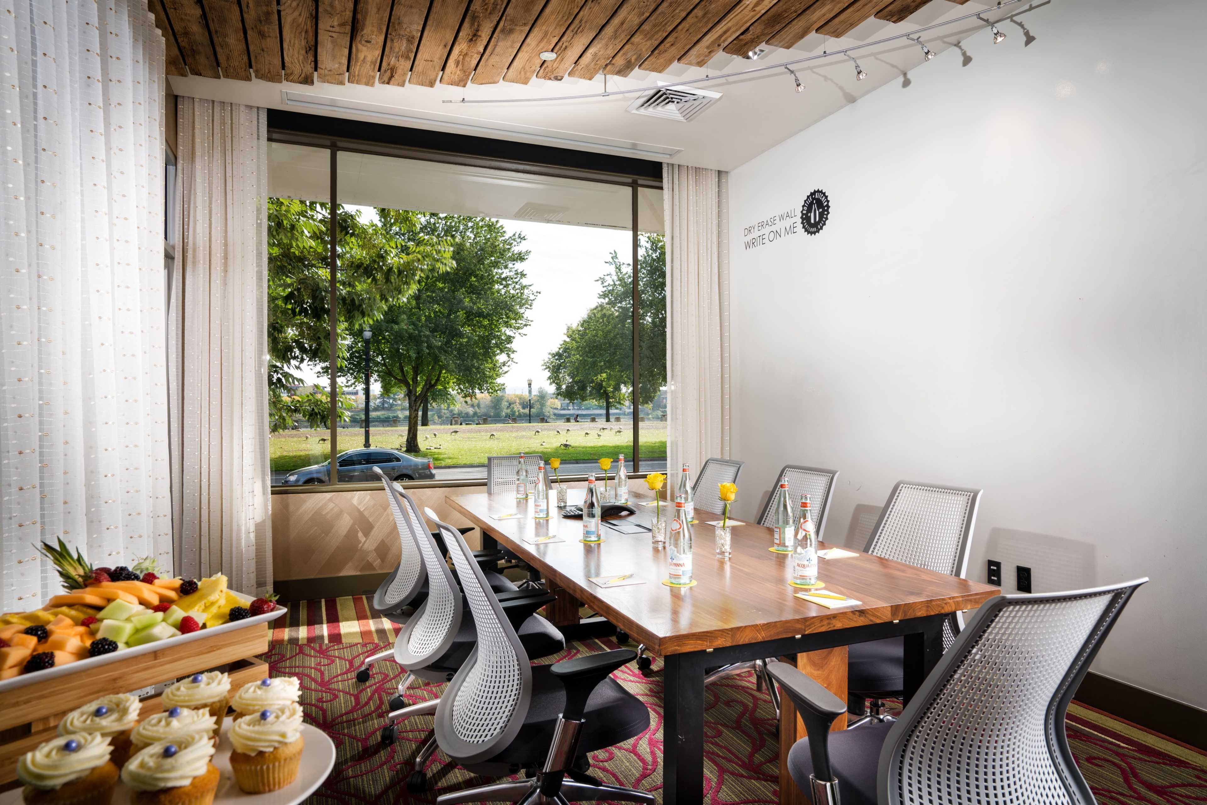 A boardroom with a wooden table, ergonomic chairs, and a view of an outdoor landscape featuring trees and a parked car.