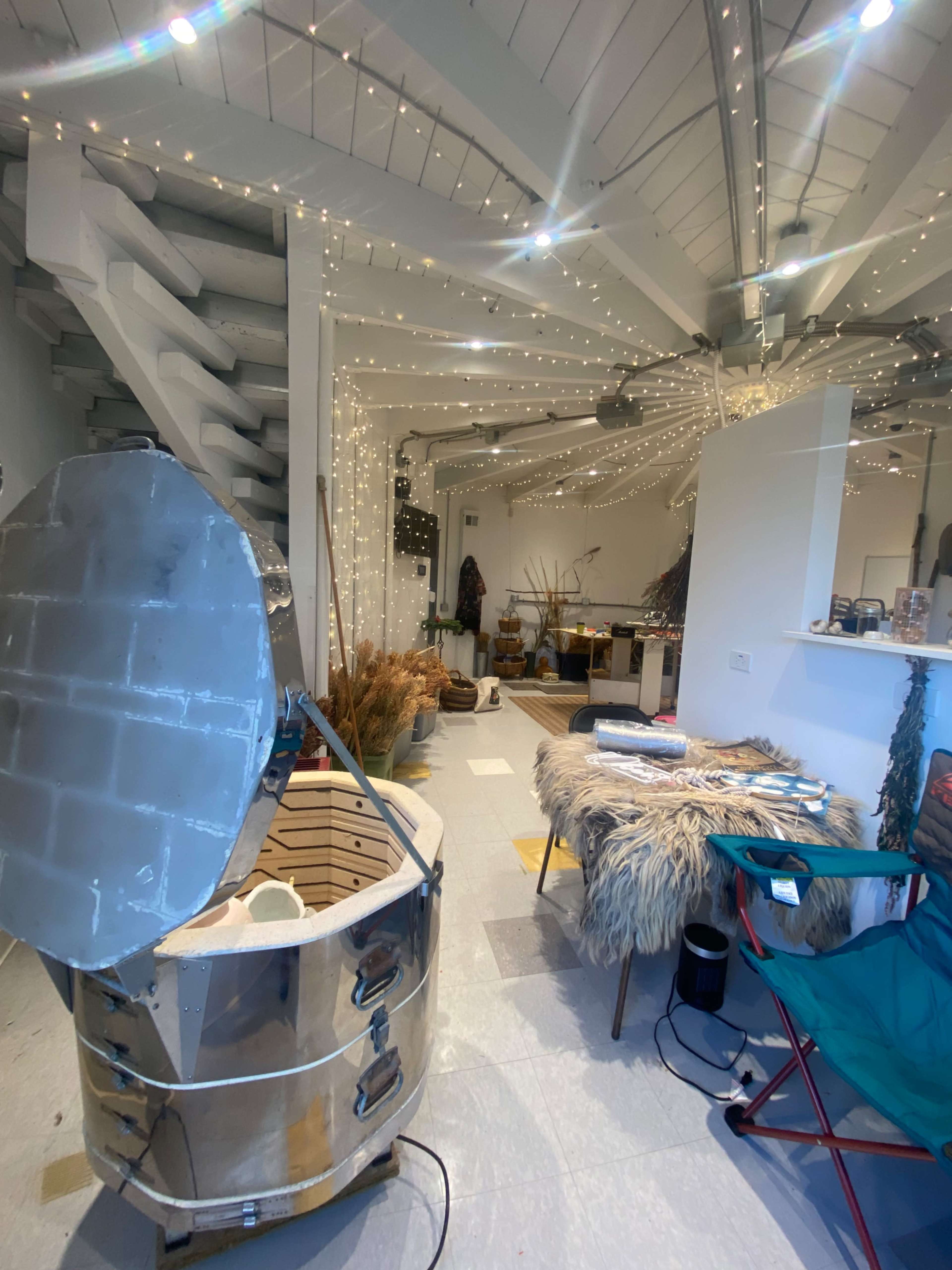 The image shows an open space with wooden beams, ambient string lights, and various decor items, including a round metallic tub and a chair.