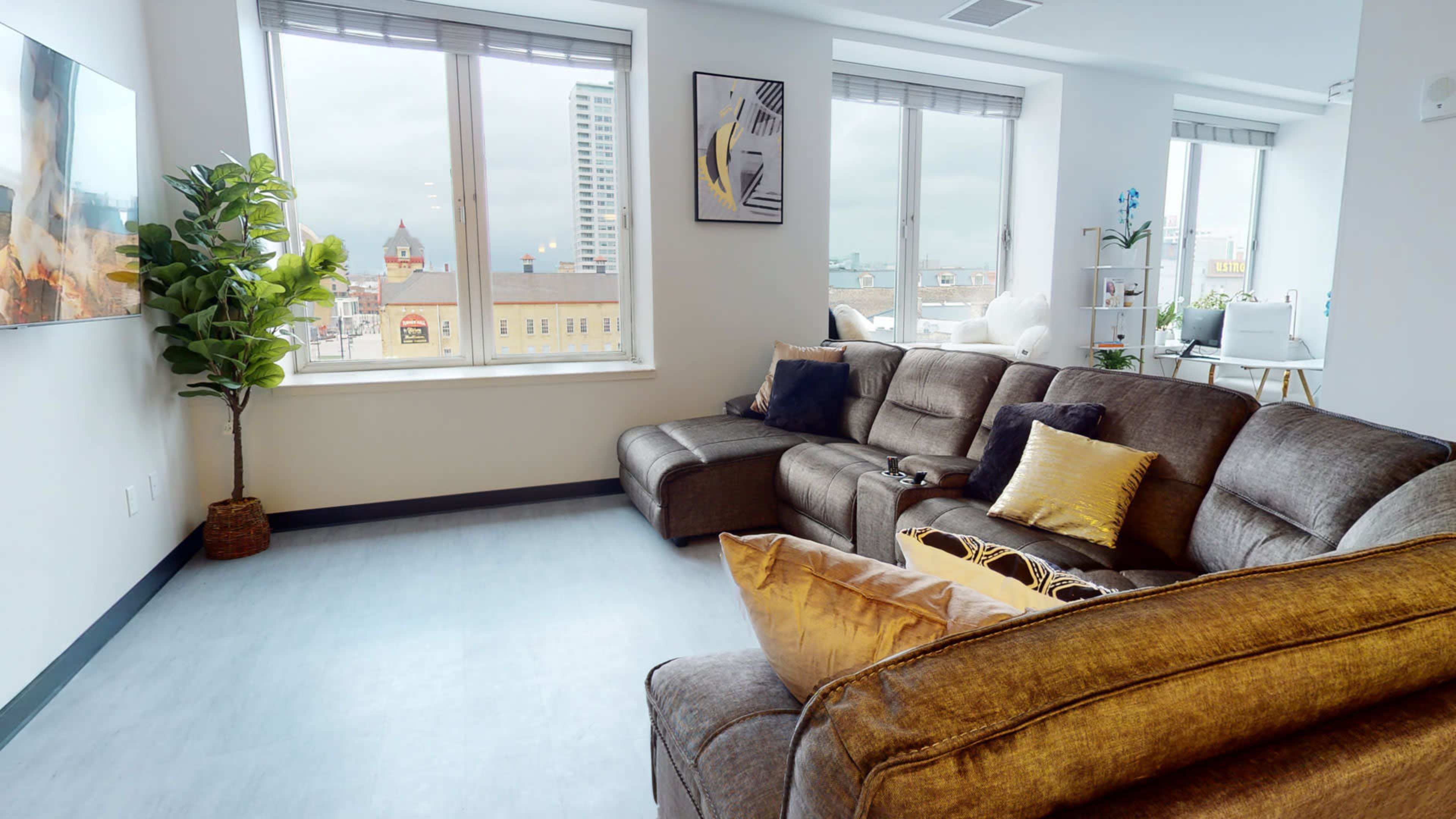 A modern living room features a sectional sofa, large windows with city views, and a potted plant.
