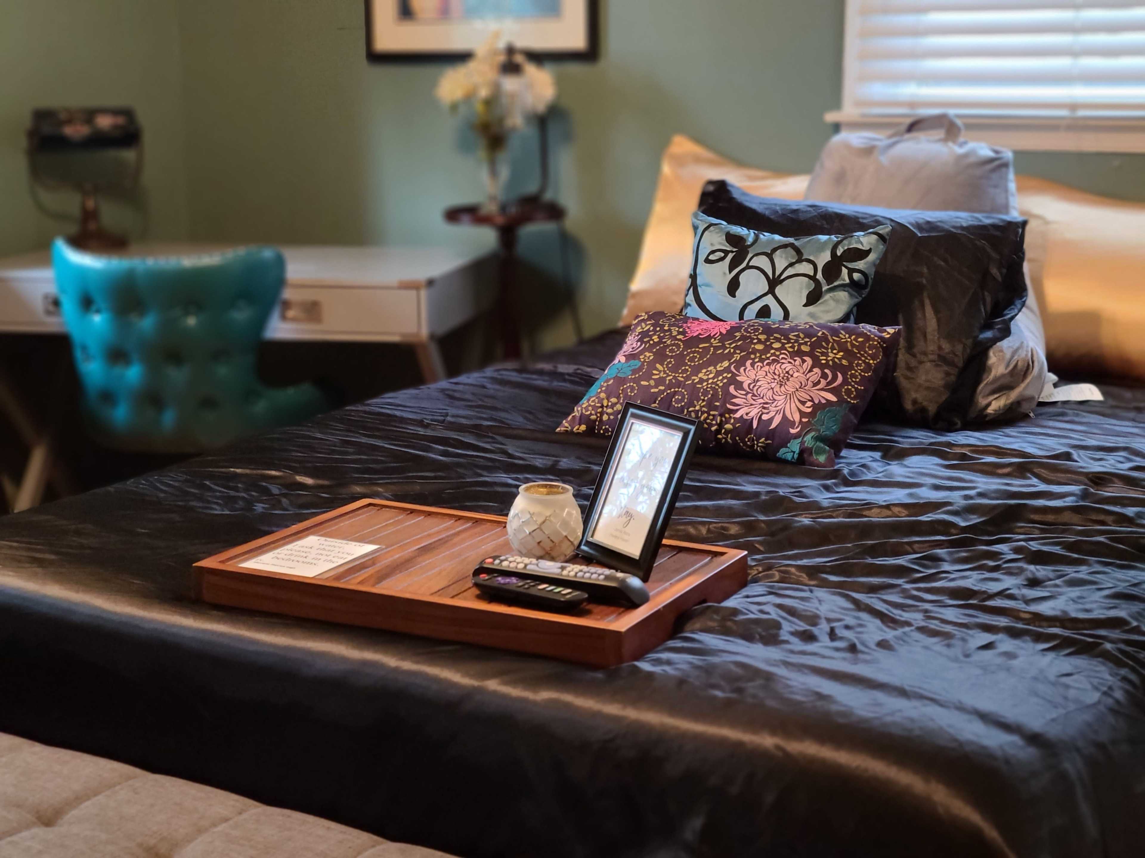 A neatly arranged bed with decorative pillows and a wooden tray holding a small vase and remote controls sits in a well-lit bedroom.