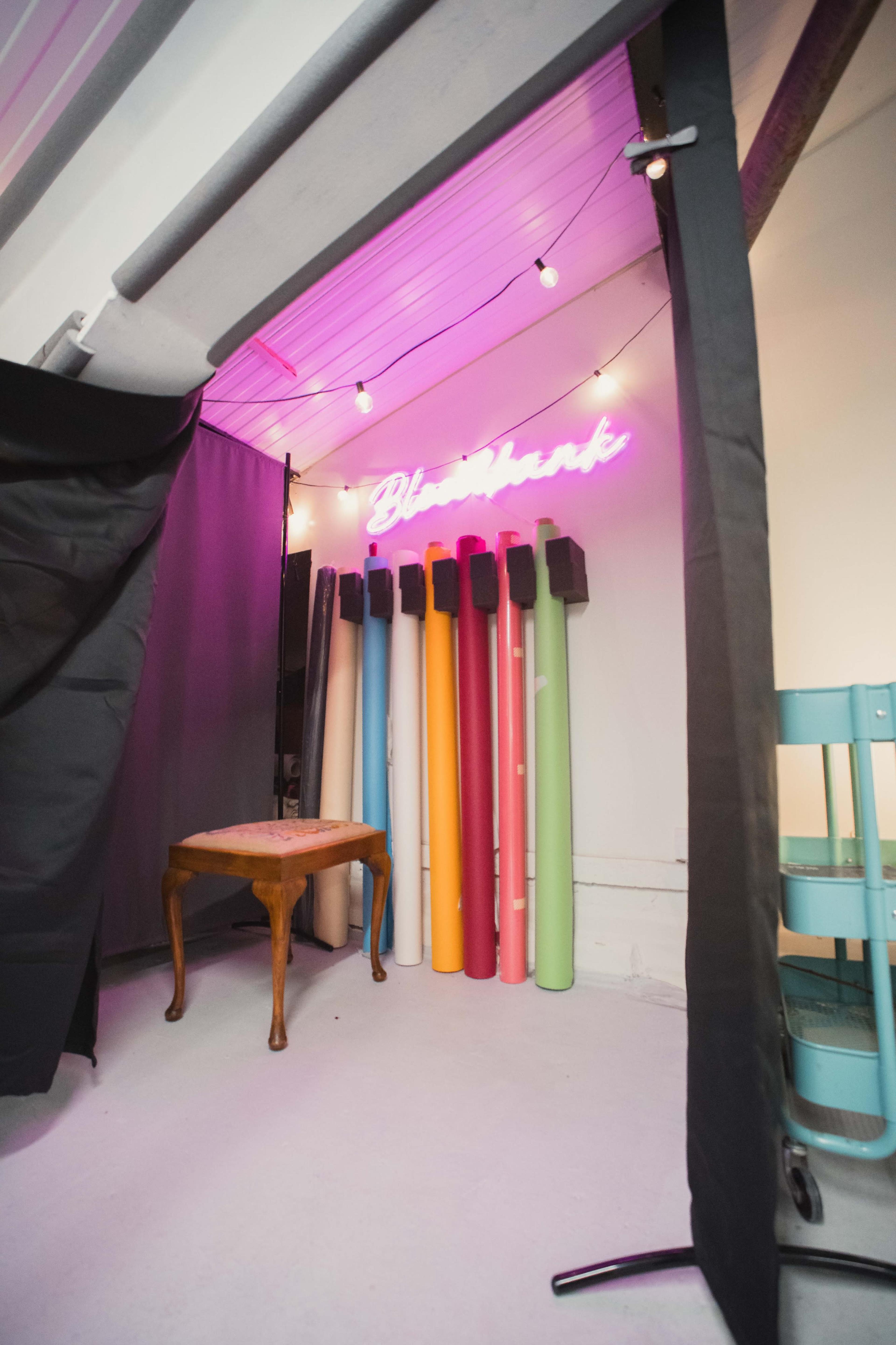 The image shows a studio space with a neon sign reading "Blastbank," a table, and several colorful paper rolls positioned against the wall.