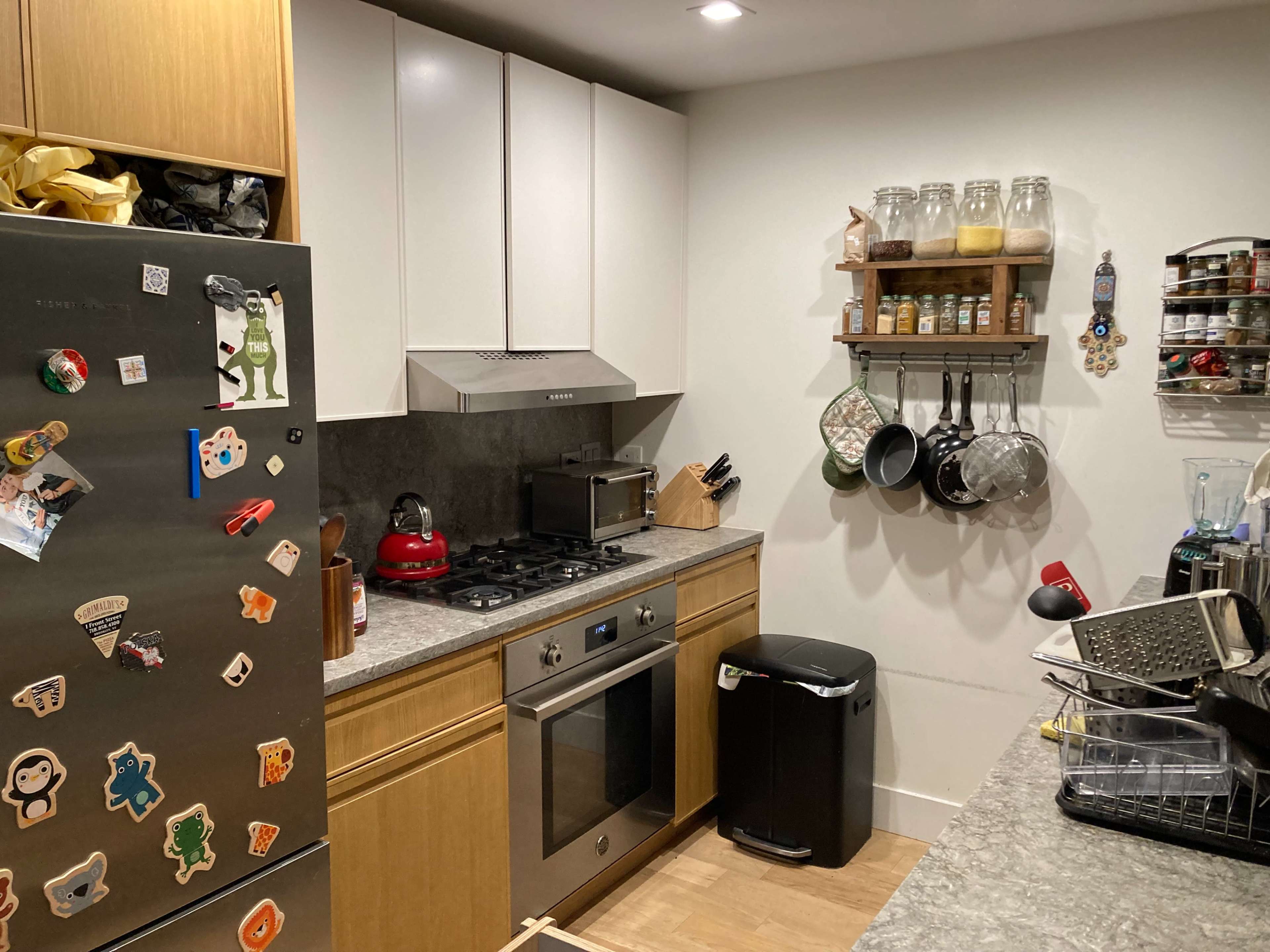 The image depicts a compact kitchen with a stainless steel refrigerator, a gas stove, and a hanging shelf displaying pots and various kitchen items.