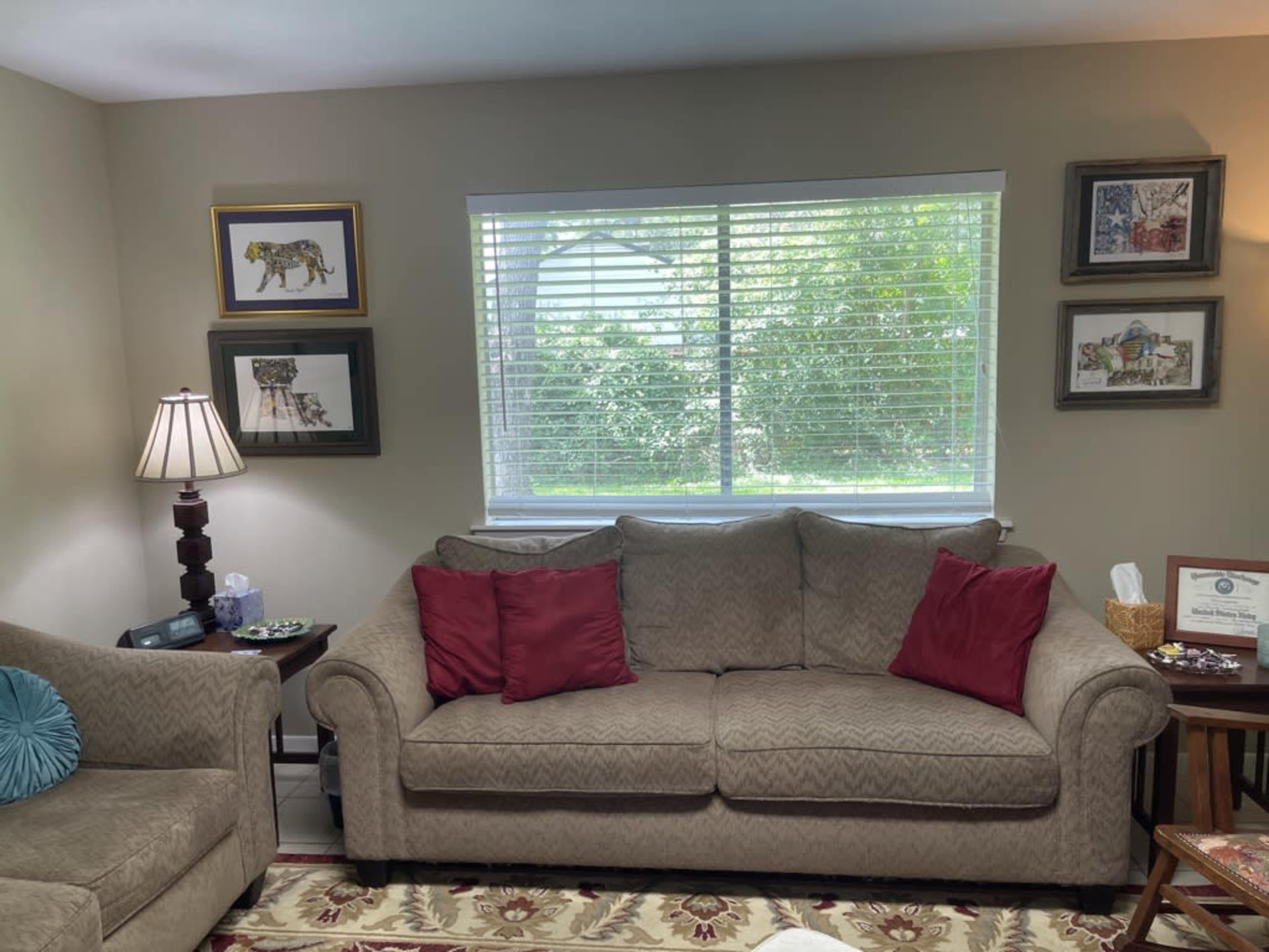 A living room featuring a beige couch with red pillows, a lamp on a side table, and framed artwork on the walls, with a window providing natural light and a view of greenery outside.