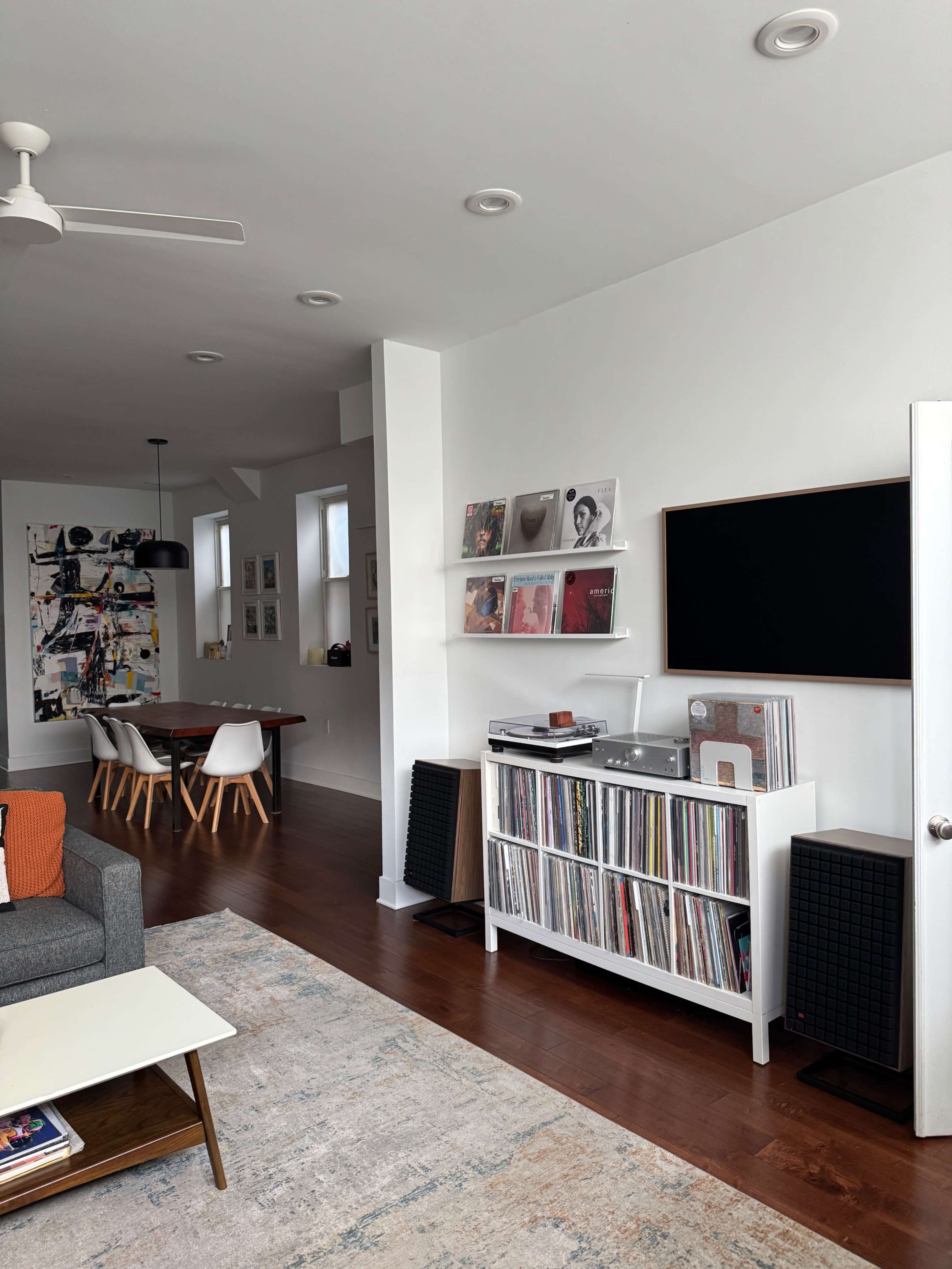 The image shows a modern living room featuring a white record shelf filled with vinyl records, a gray sofa, and a dining area with a table and chairs in the background.