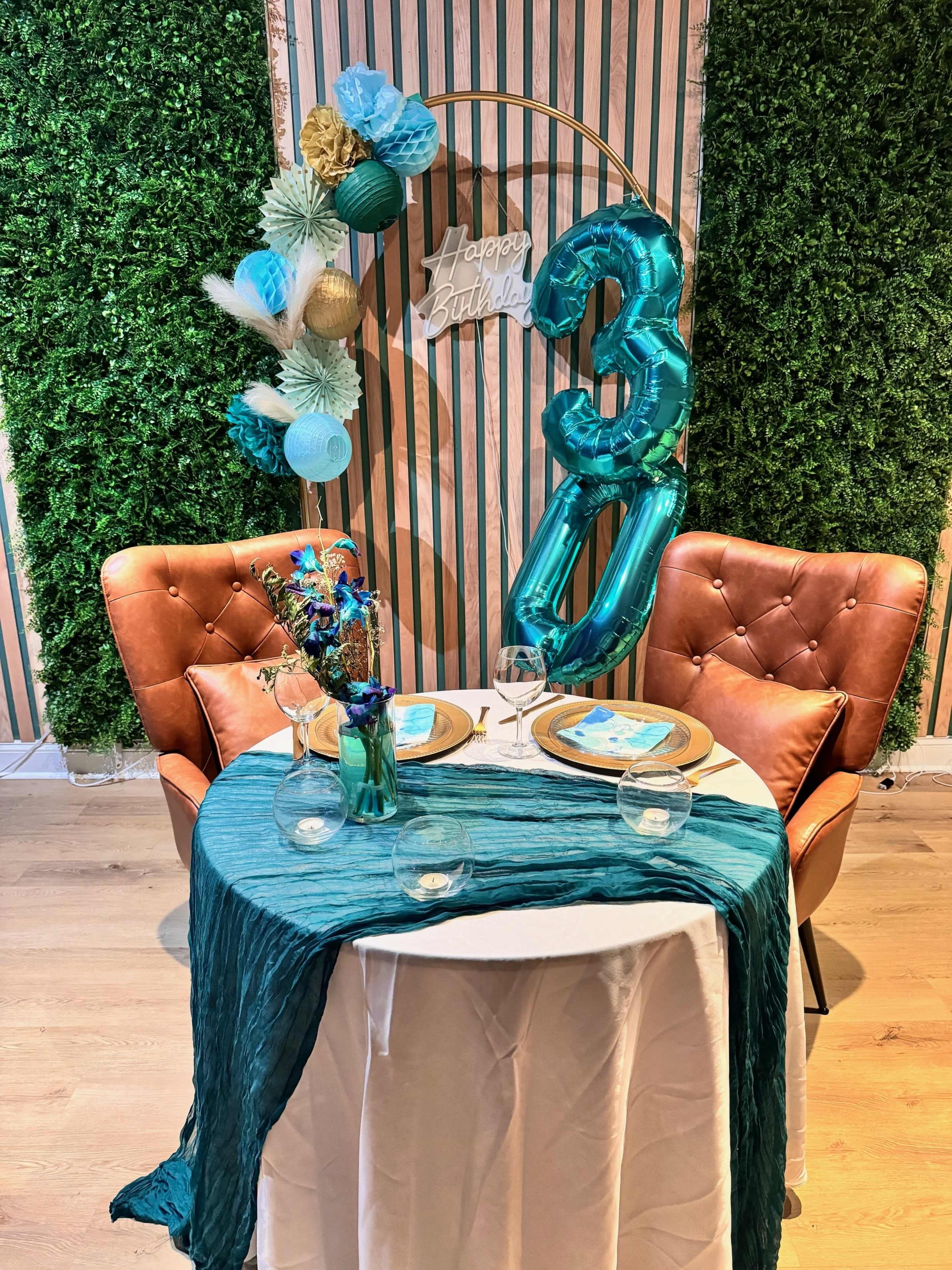 The image shows a decorated dining setup featuring a round table with a teal tablecloth, two brown chairs, a floral centerpiece, and "Happy Birthday" balloons in the shape of the number 30 against a backdrop of greenery and vertical stripes.