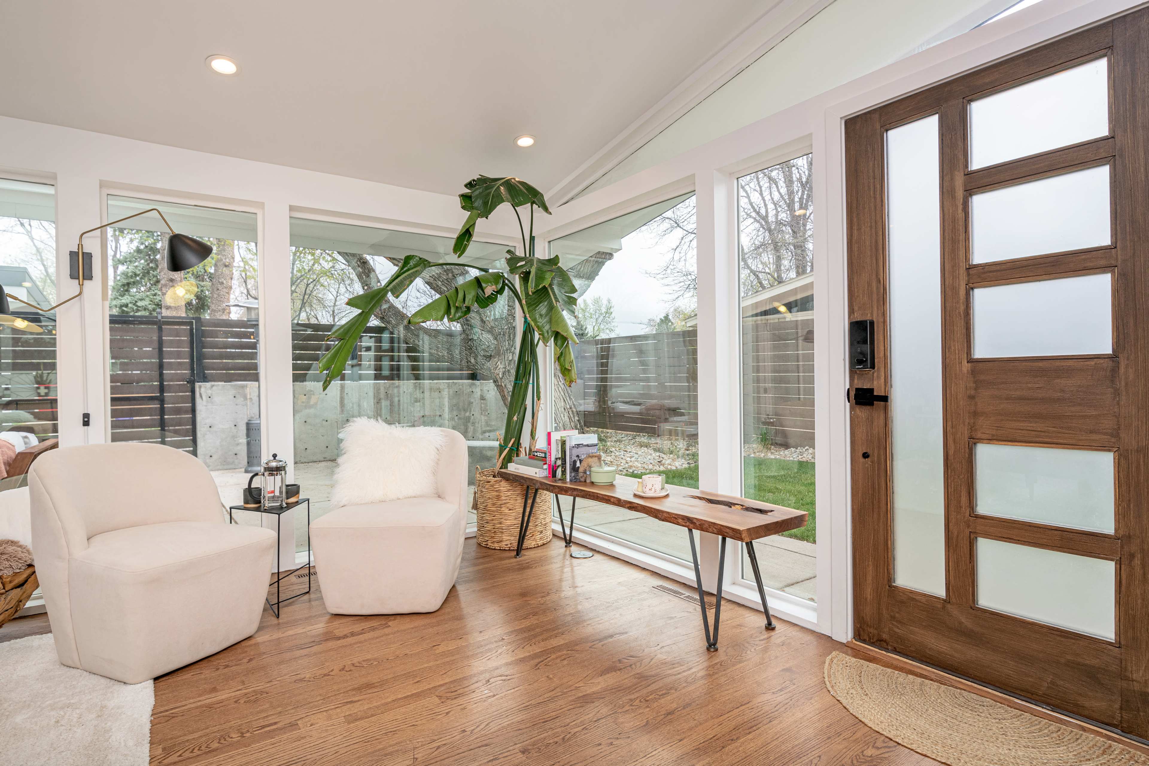 A bright corner of a room features two chairs, a wooden table with items on it, and large windows that overlook a garden area.
