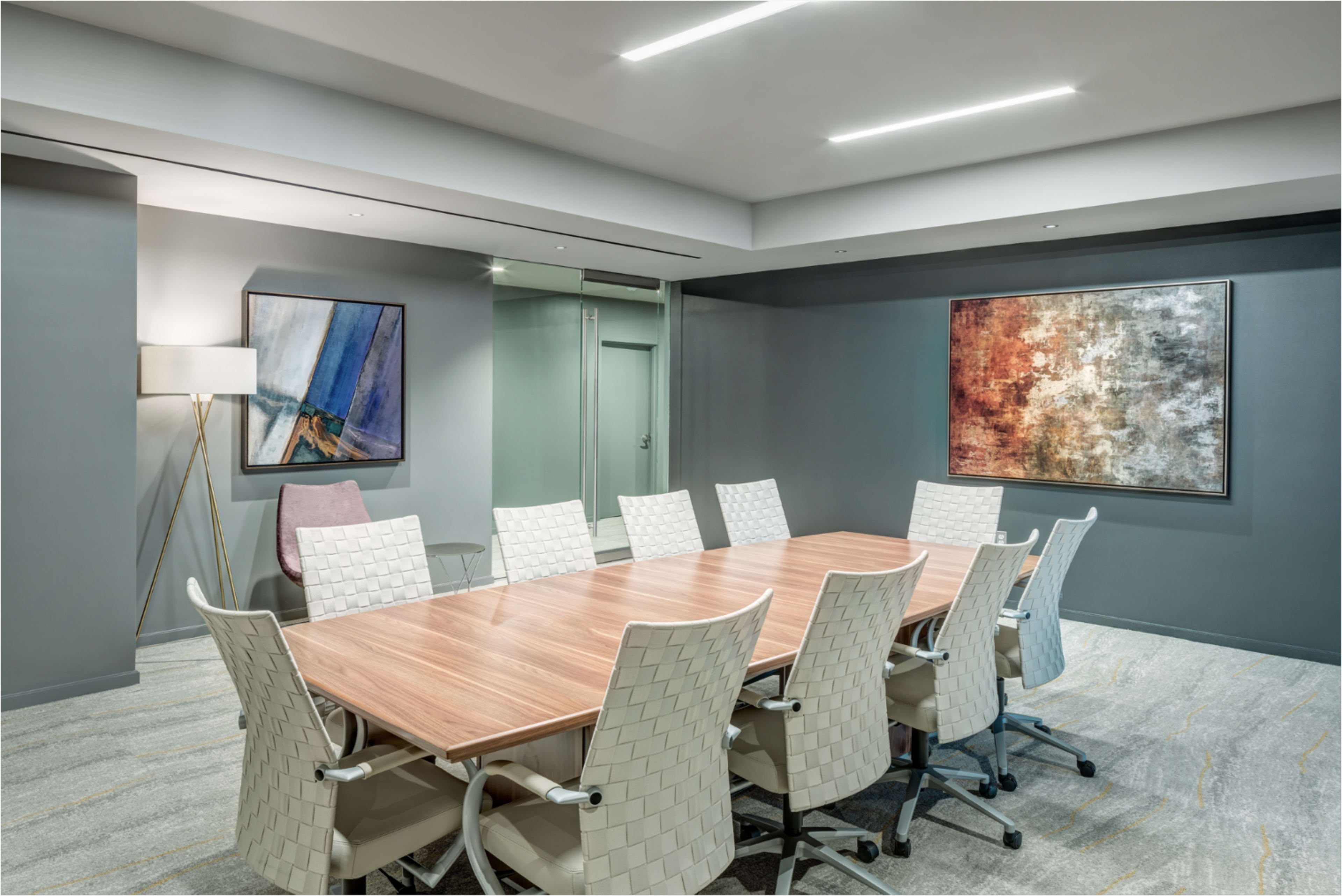 A modern conference room features a long wooden table surrounded by twelve white, woven chairs, with abstract artwork hanging on the walls.