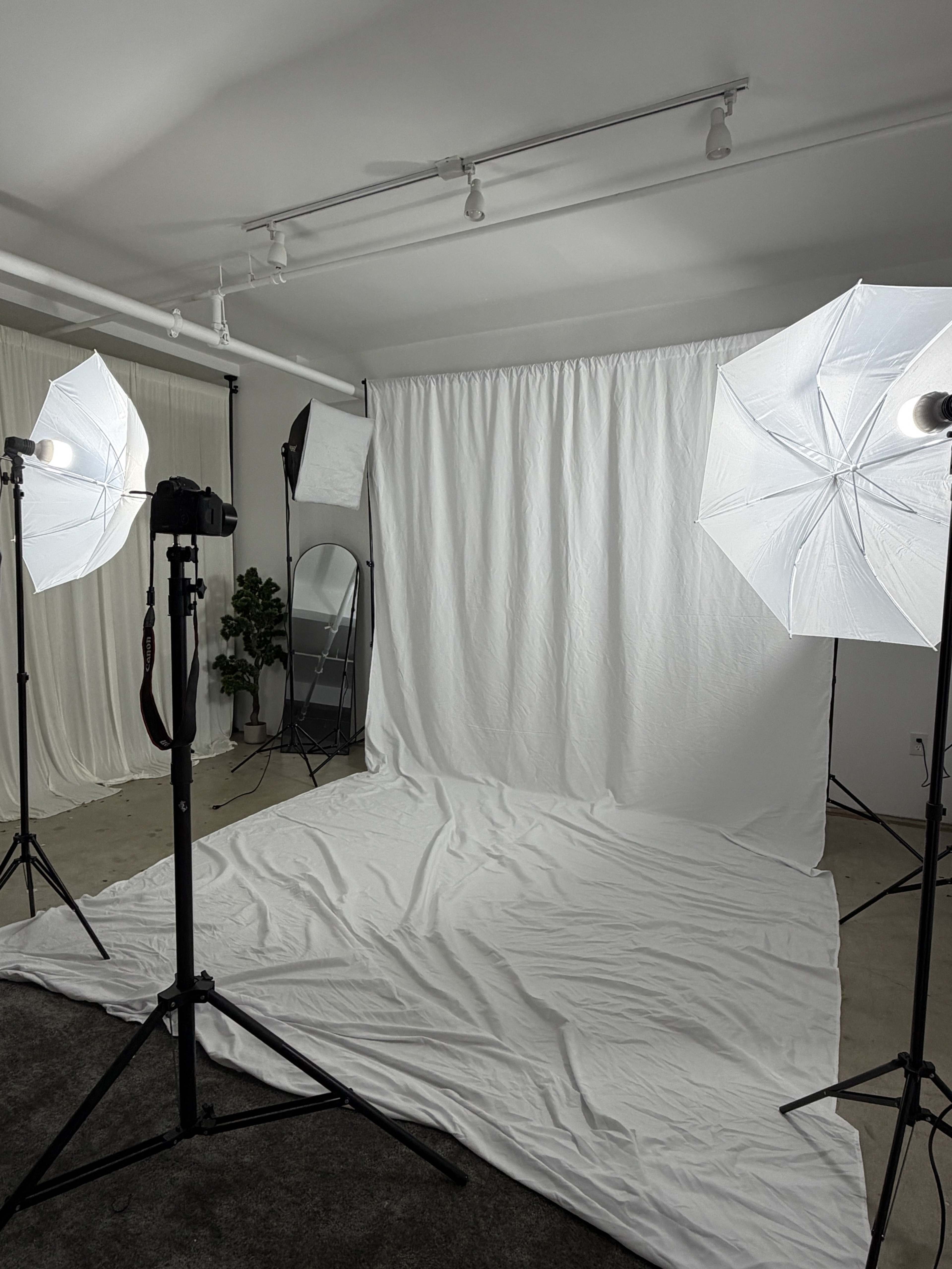 The image shows a photography studio setup with white backdrops, umbrella softboxes for lighting, and a mirror placed in the corner.