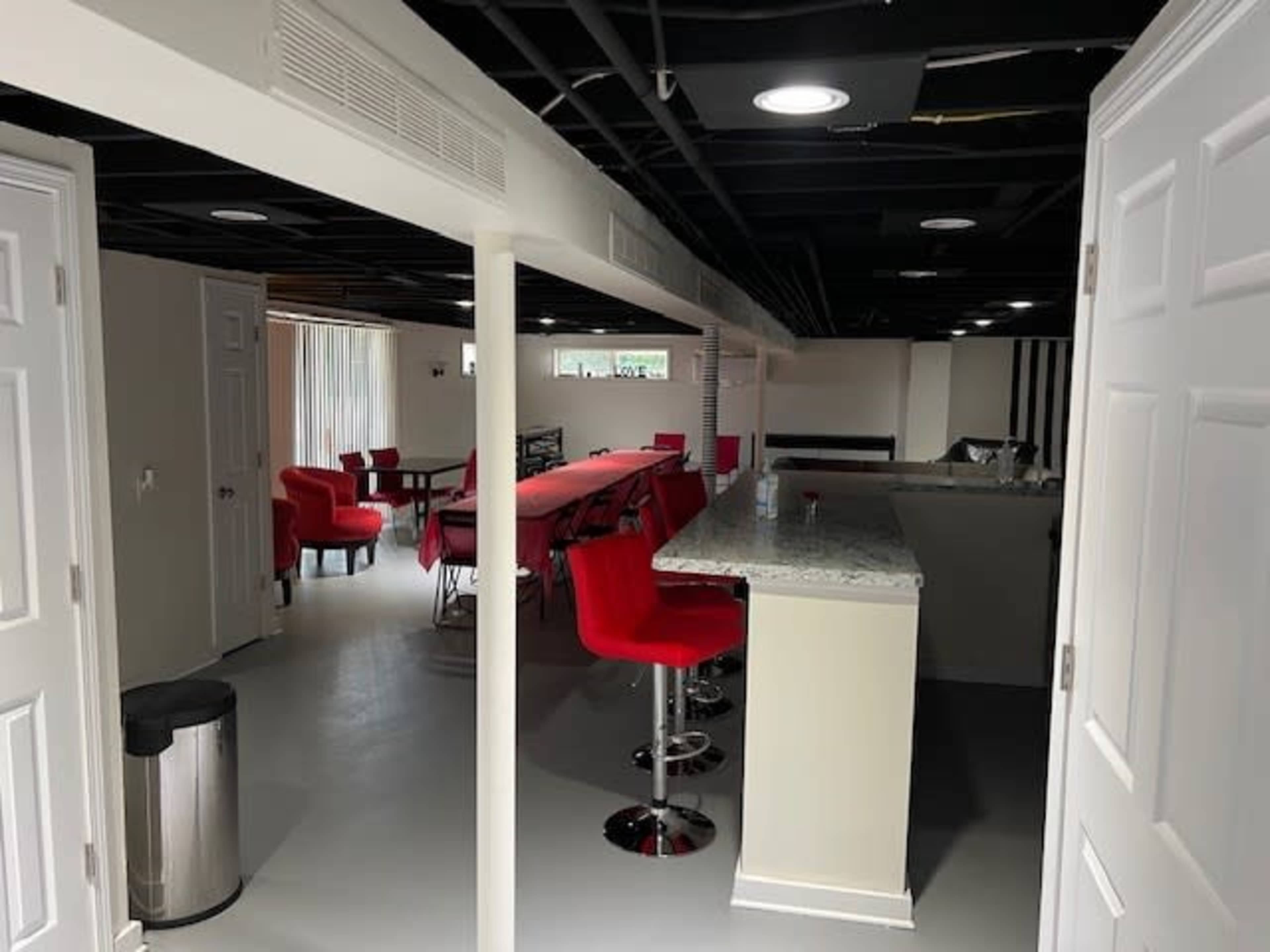 A spacious basement area with a bar counter, red stools, and a long dining table surrounded by red chairs.