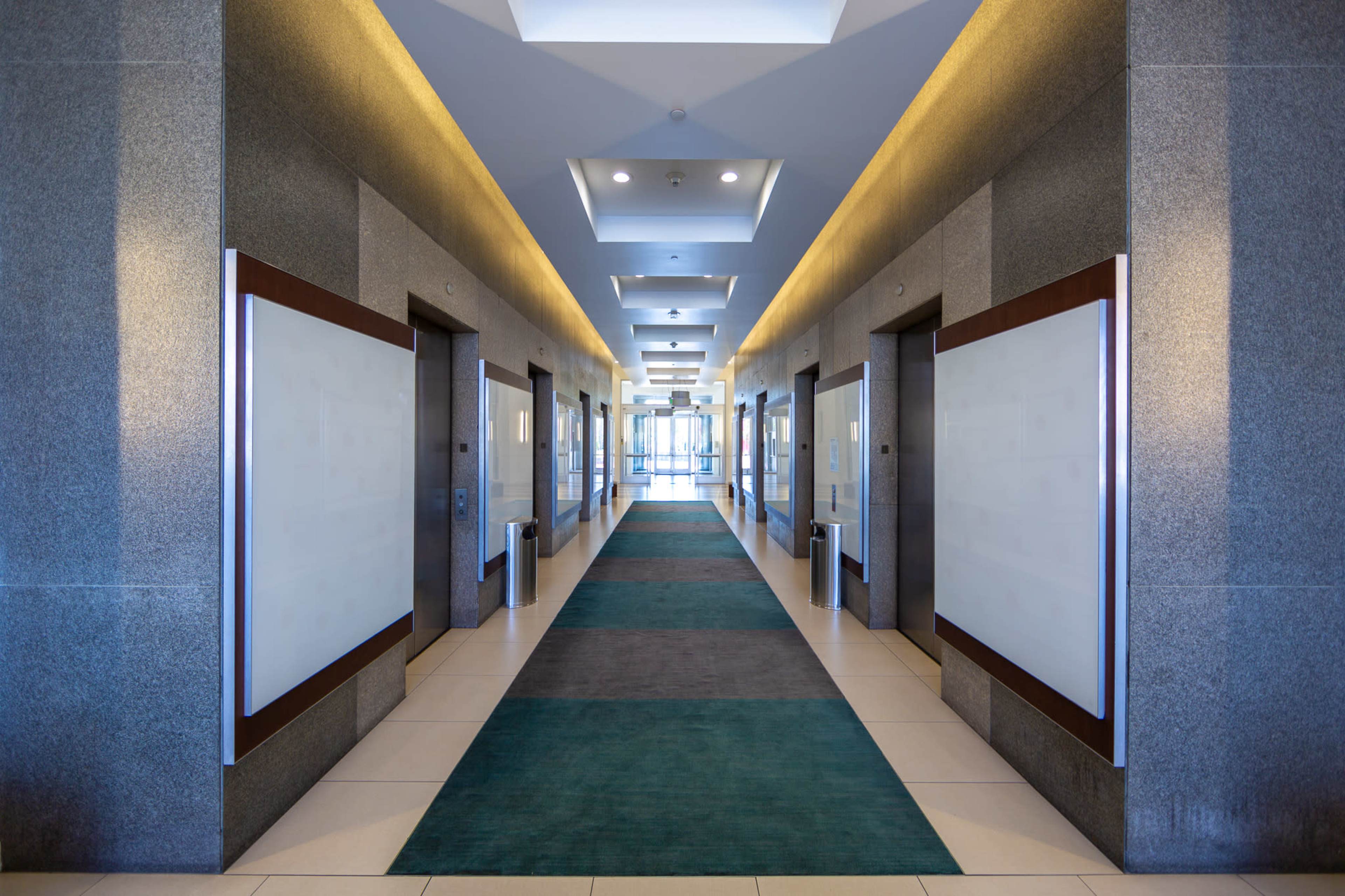 The image shows a long corridor with gray walls, a central green carpet, and glass doors at the far end.
