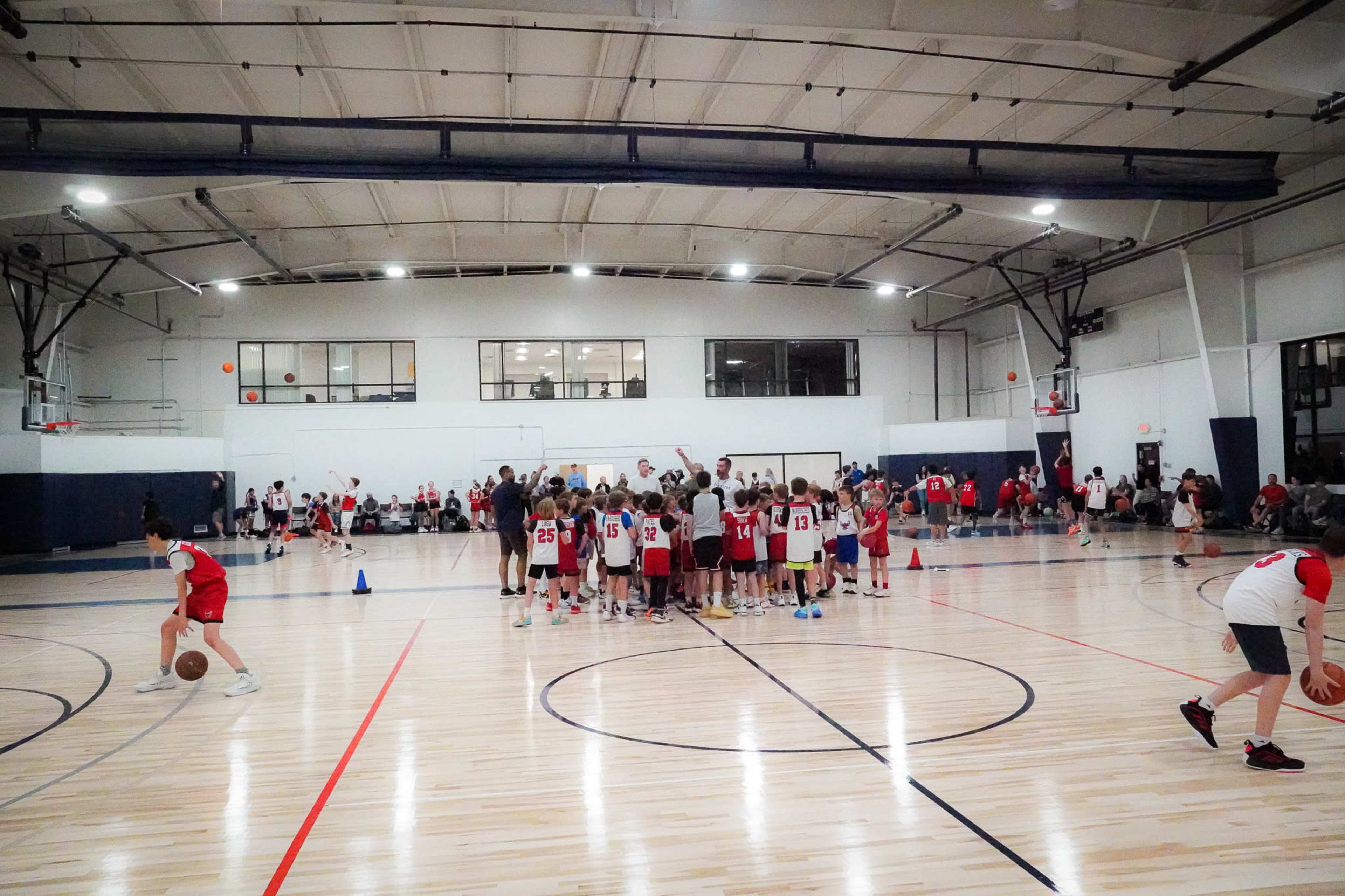 A group of young basketball players in red and white jerseys gathers together in a gymnasium, while others practice shooting on the court.