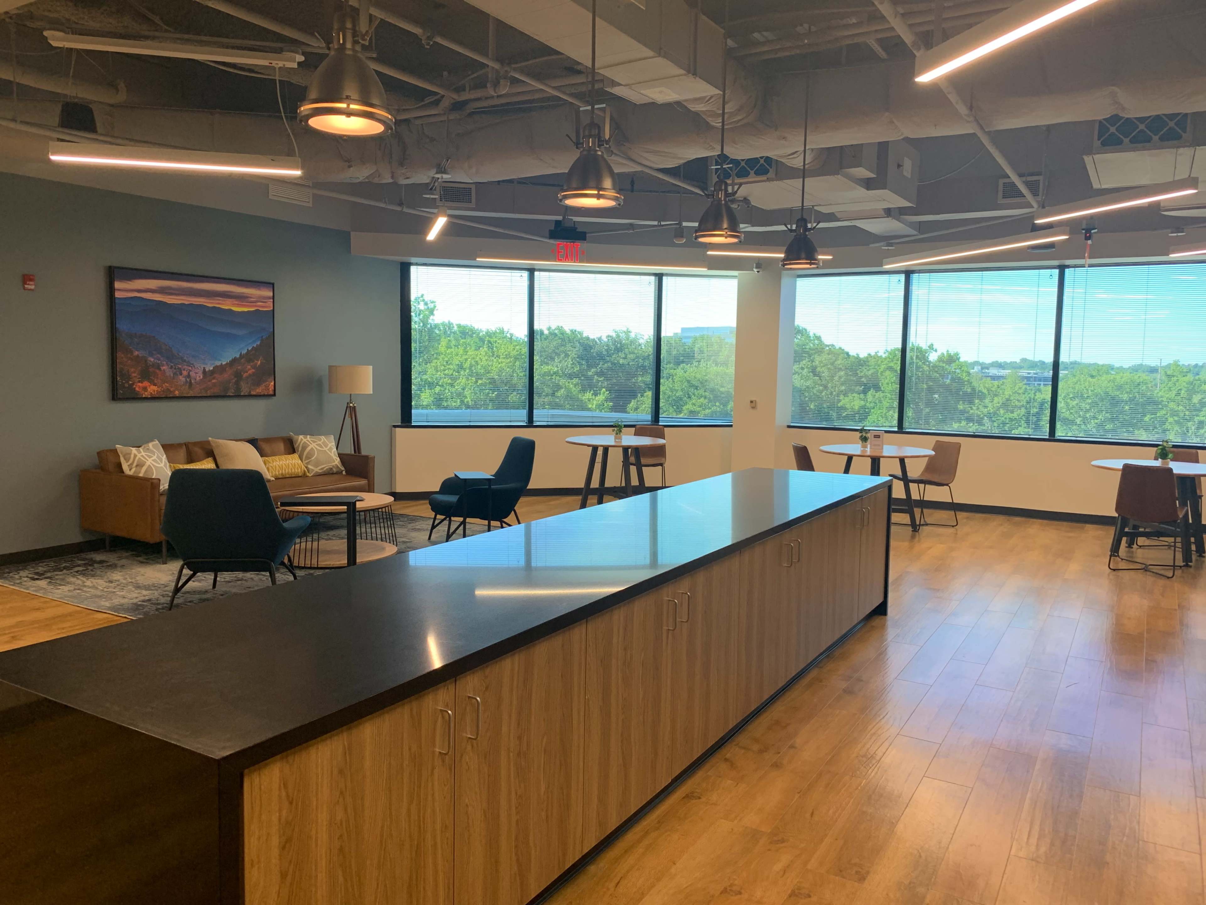 The image shows a modern office lounge area with a central dark countertop, seating options, and large windows overlooking greenery.