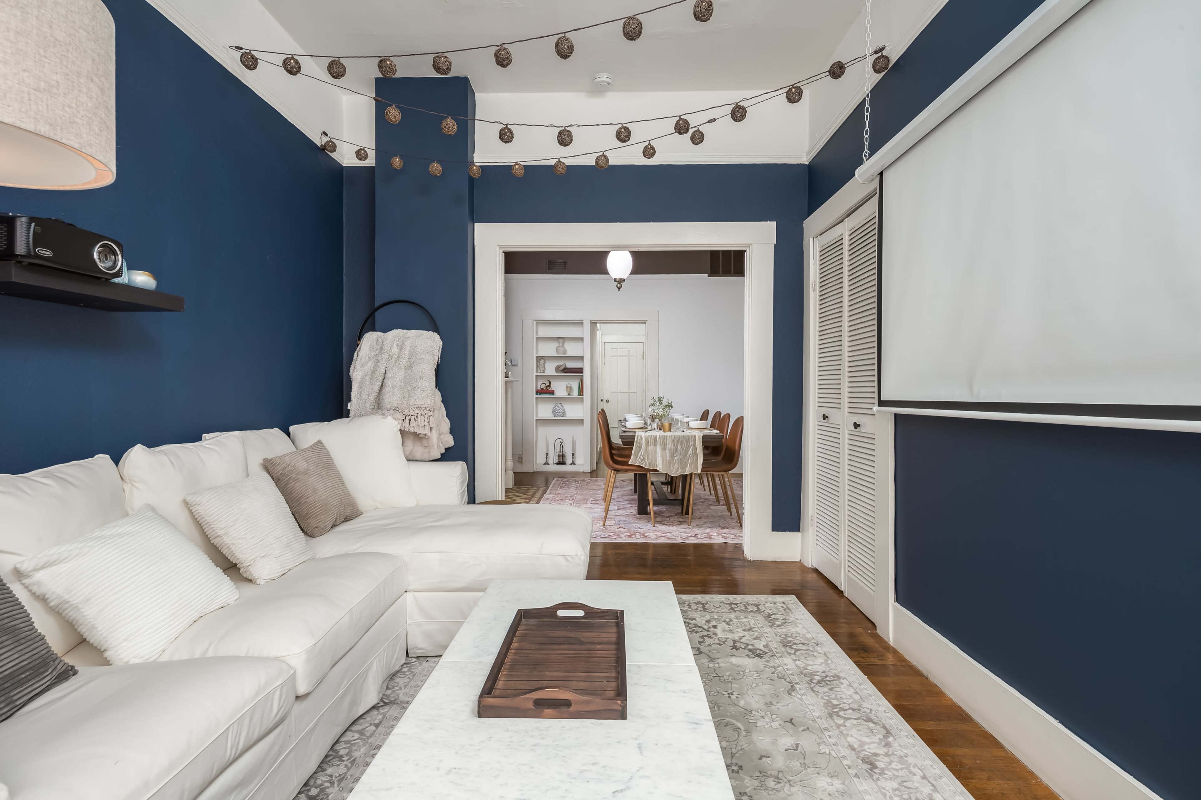 A cozy living room with a white sectional sofa, a marble coffee table, and blue walls, leading to a dining area in the background.