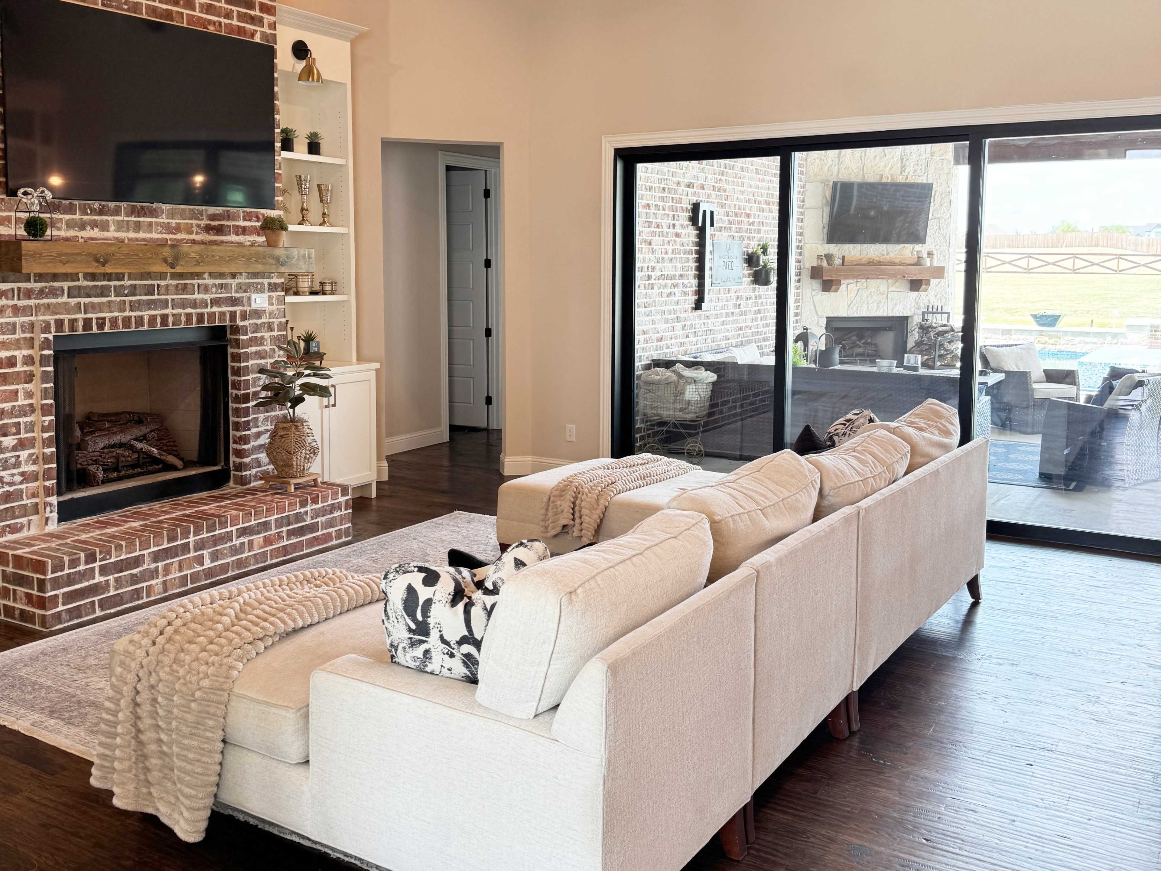 The image shows a living room featuring a beige sectional sofa facing a brick fireplace, with large glass doors leading to an outdoor patio area.