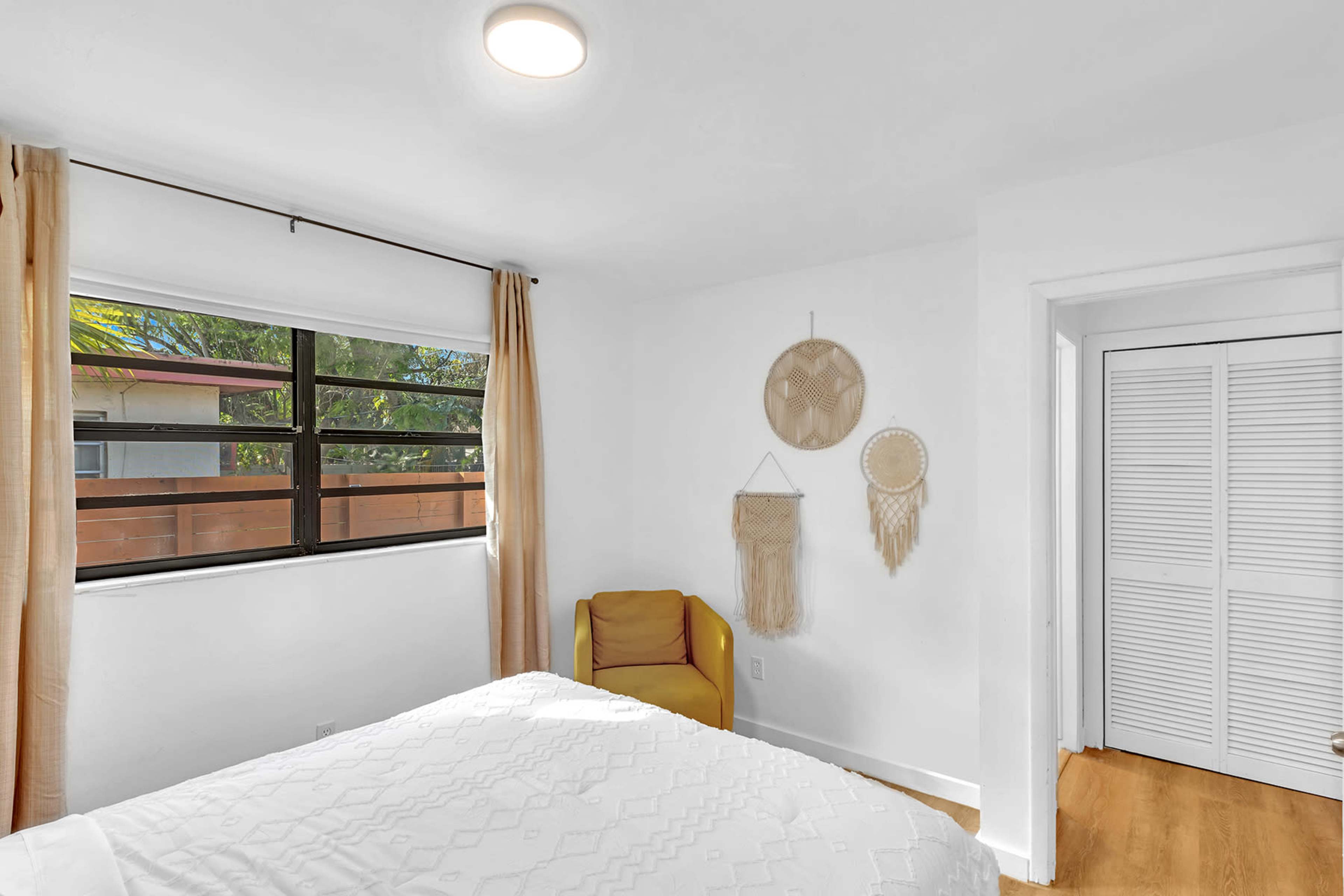 A bright bedroom featuring a bed with a white cover, a yellow chair, and wall hangings next to a large window.
