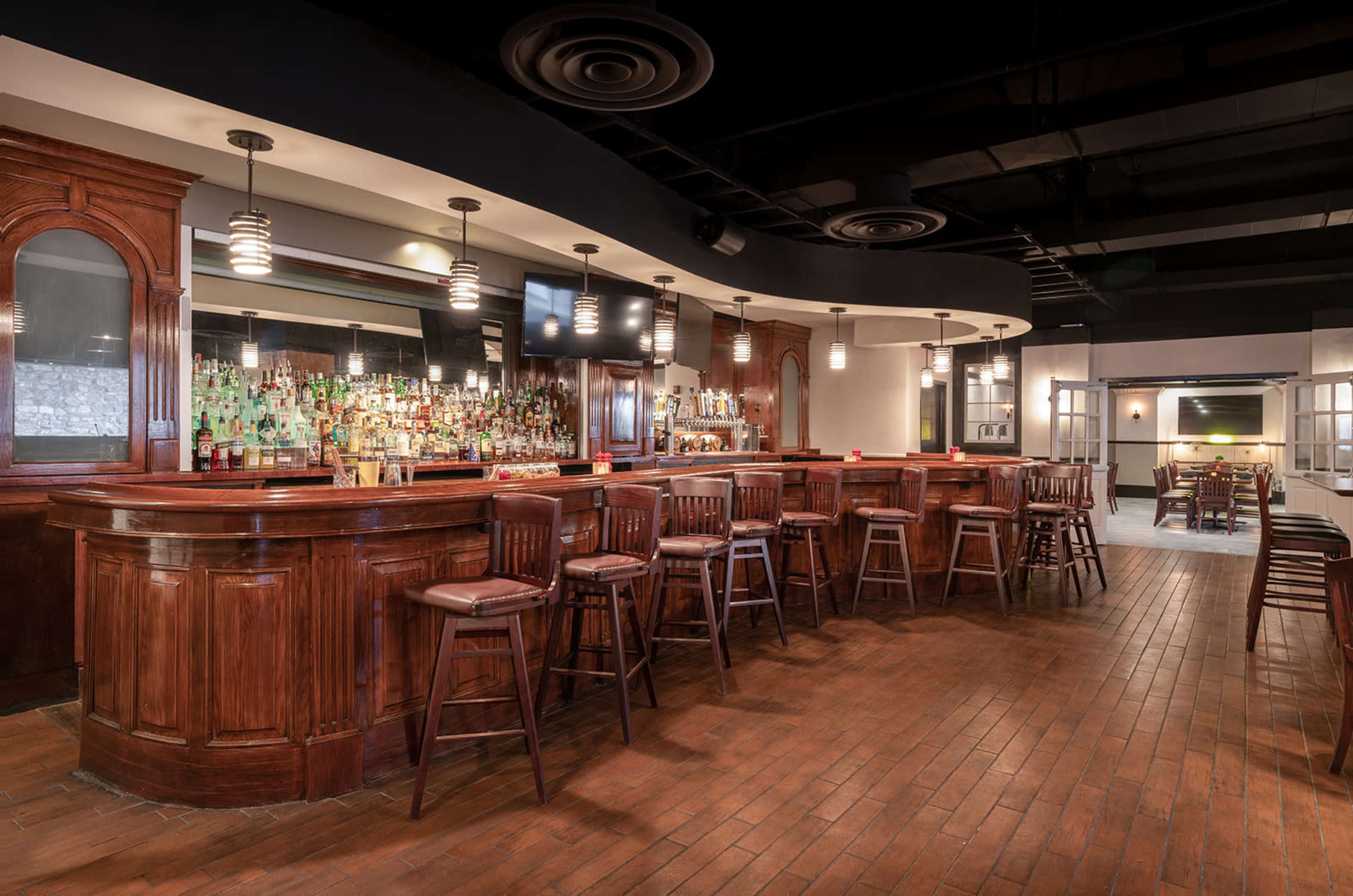 A wooden bar area with high stools, a mirrored back wall filled with bottles, and pendant lighting in a modern interior space.