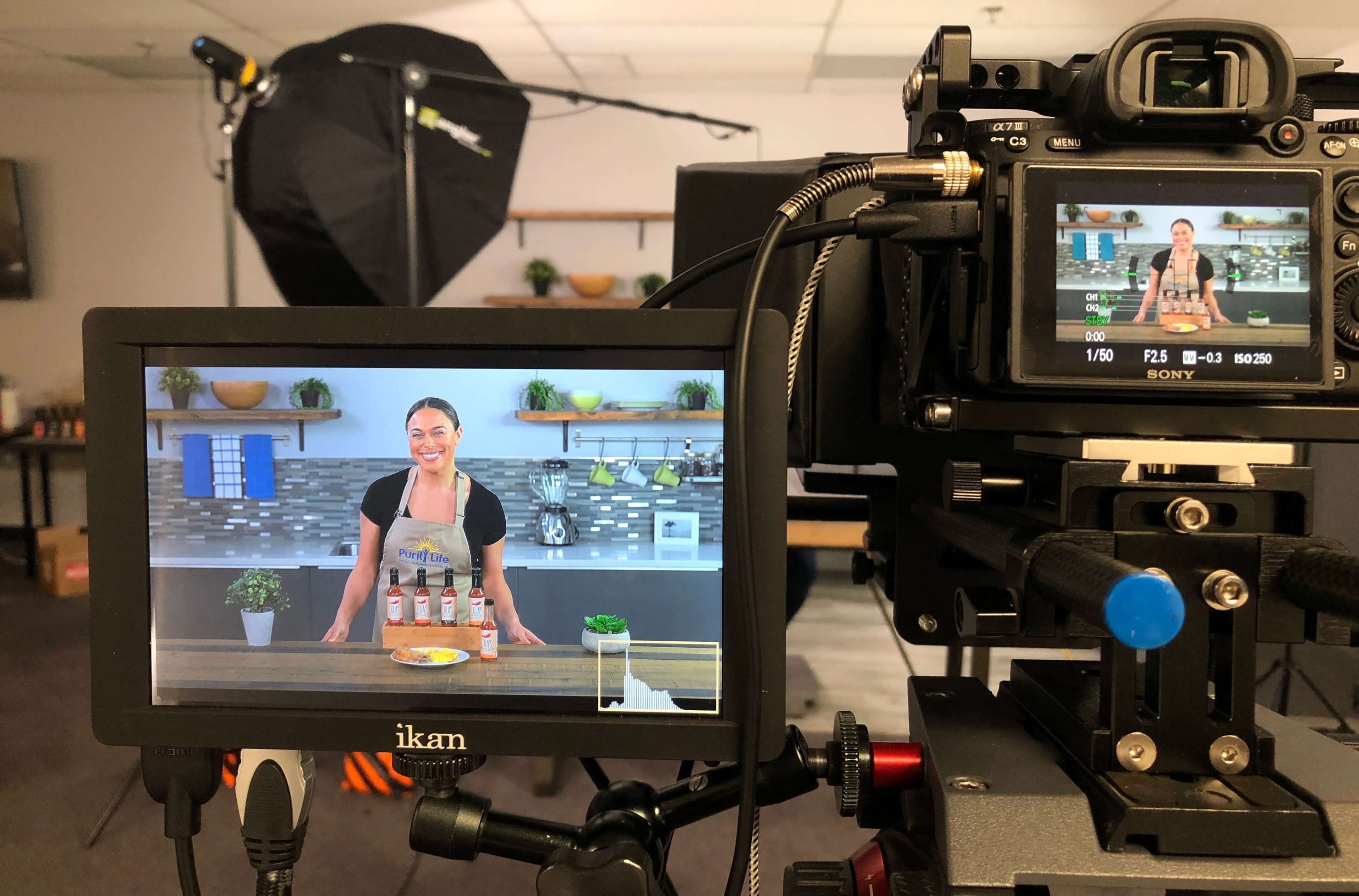 A woman stands behind a wooden table displaying several bottles of sauce in a well-lit kitchen setup, while being filmed by a camera.