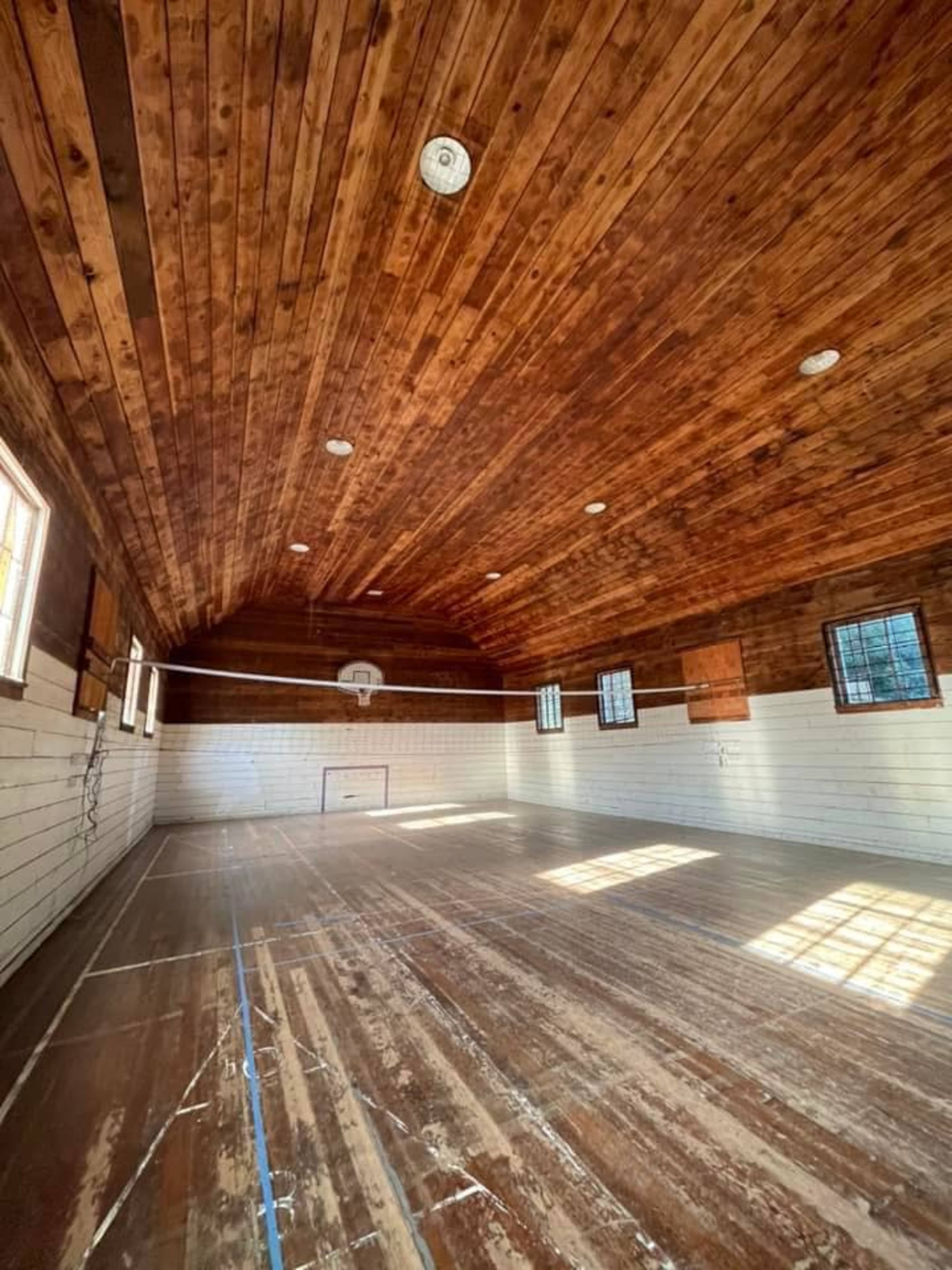 An empty wooden gymnasium with a high ceiling, basketball hoop, and natural light coming through the windows.