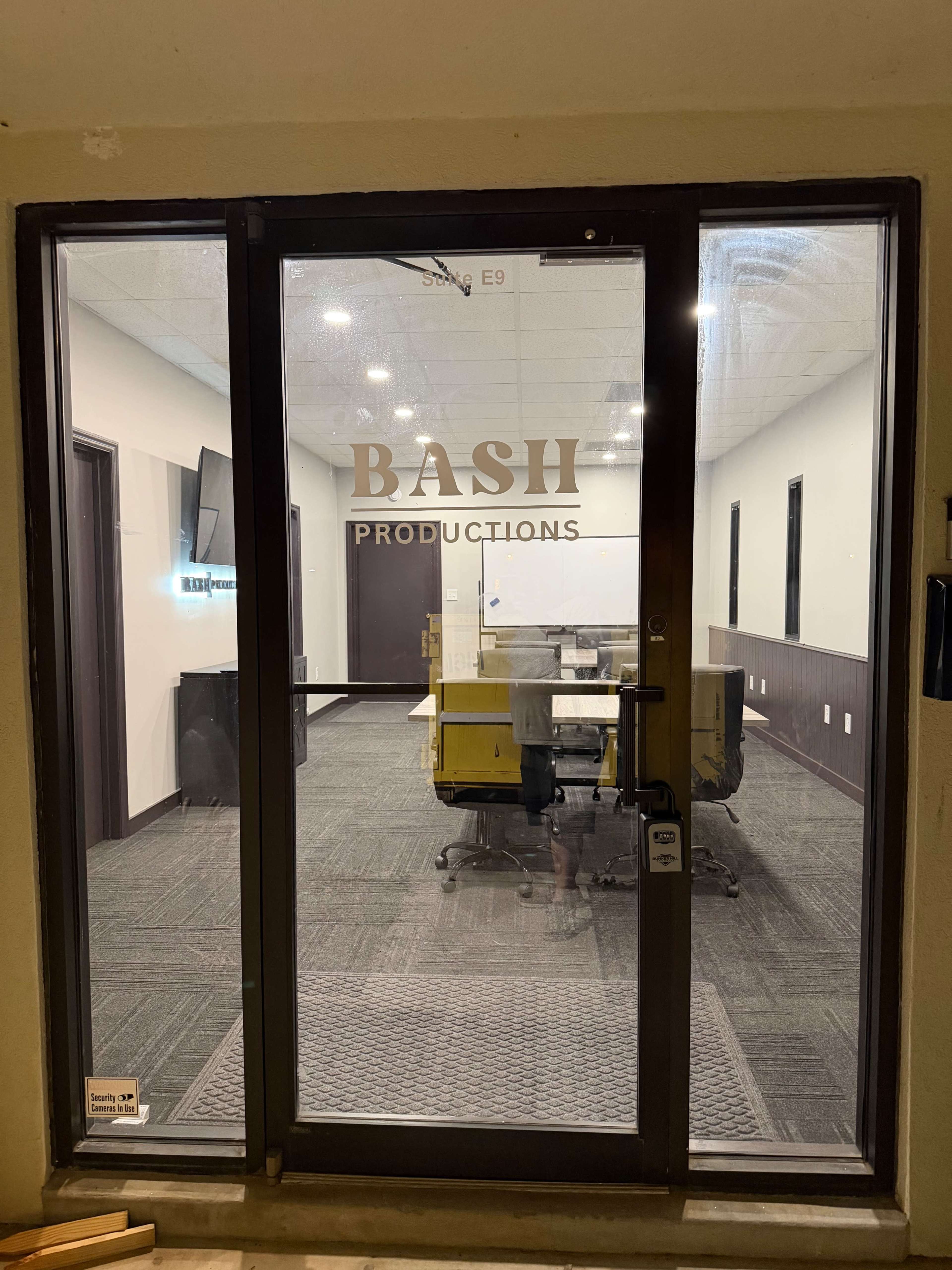 The image shows a glass door with the text "BASH PRODUCTIONS" on it, leading into an office space furnished with yellow chairs and a whiteboard.