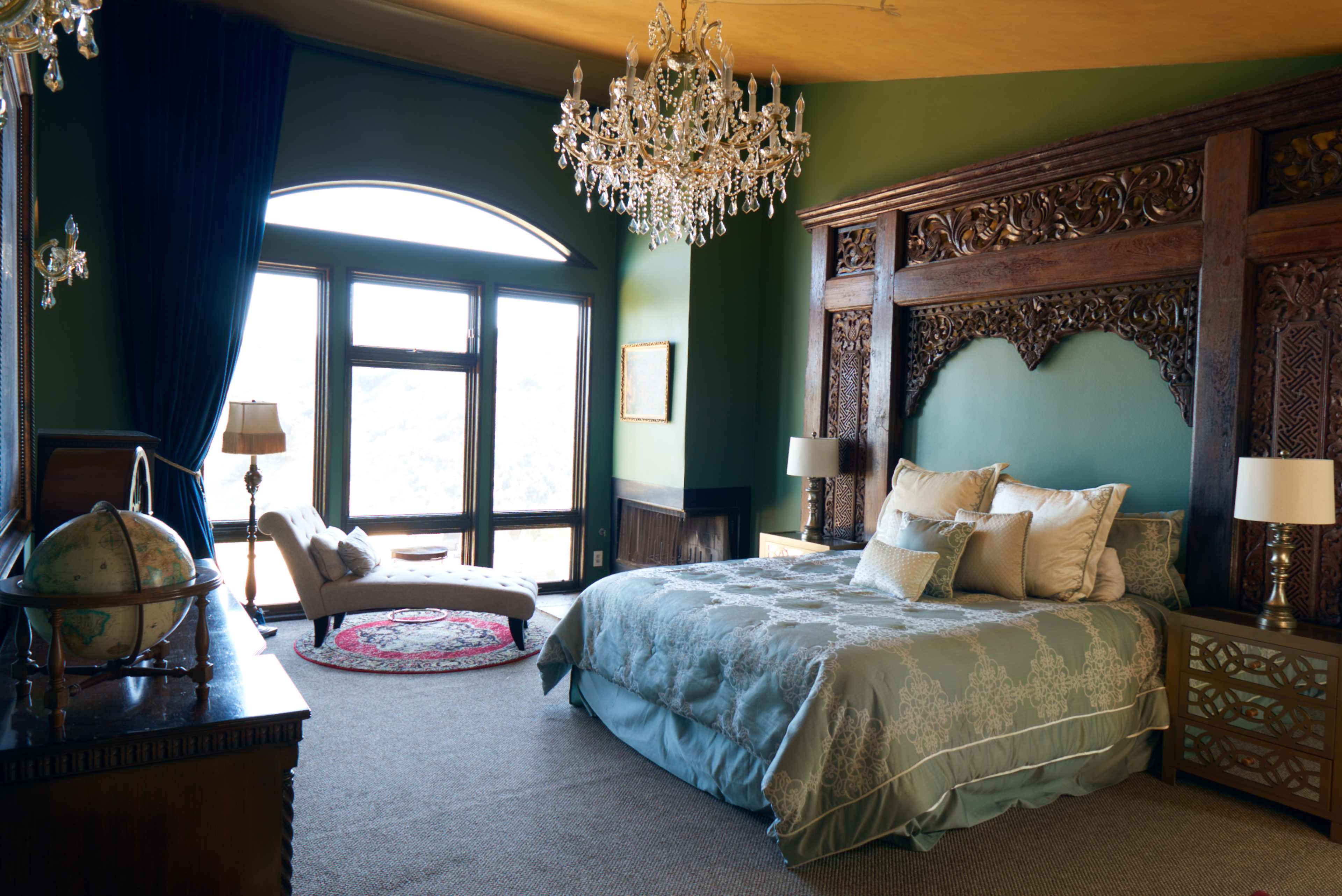 The room features a king-size bed with a decorative headboard, a crystal chandelier, and large windows overlooking the landscape.