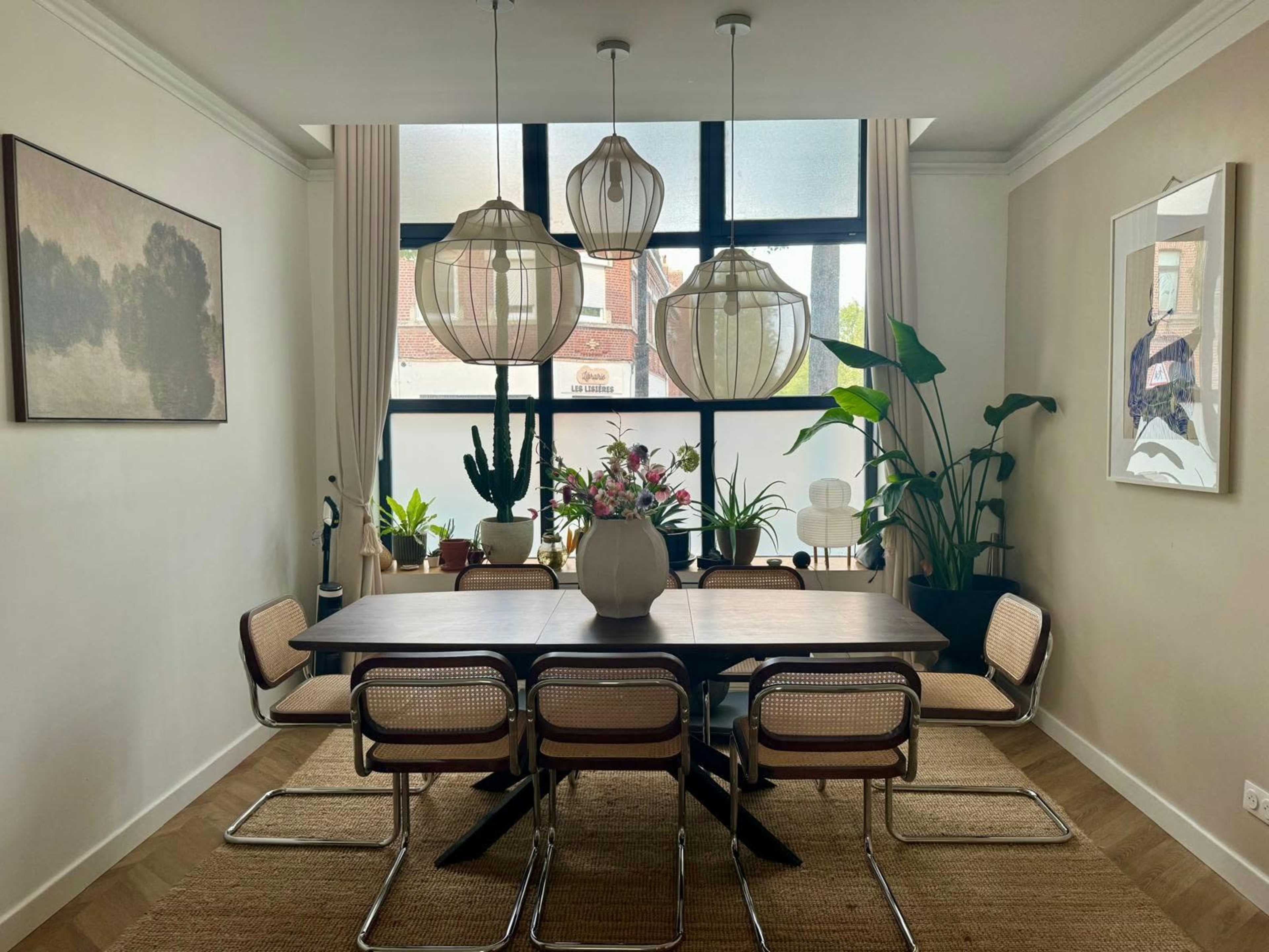 The image shows a stylish dining area with a large wooden table surrounded by chairs, illuminated by pendant lights, and decorated with plants and artwork on the walls.