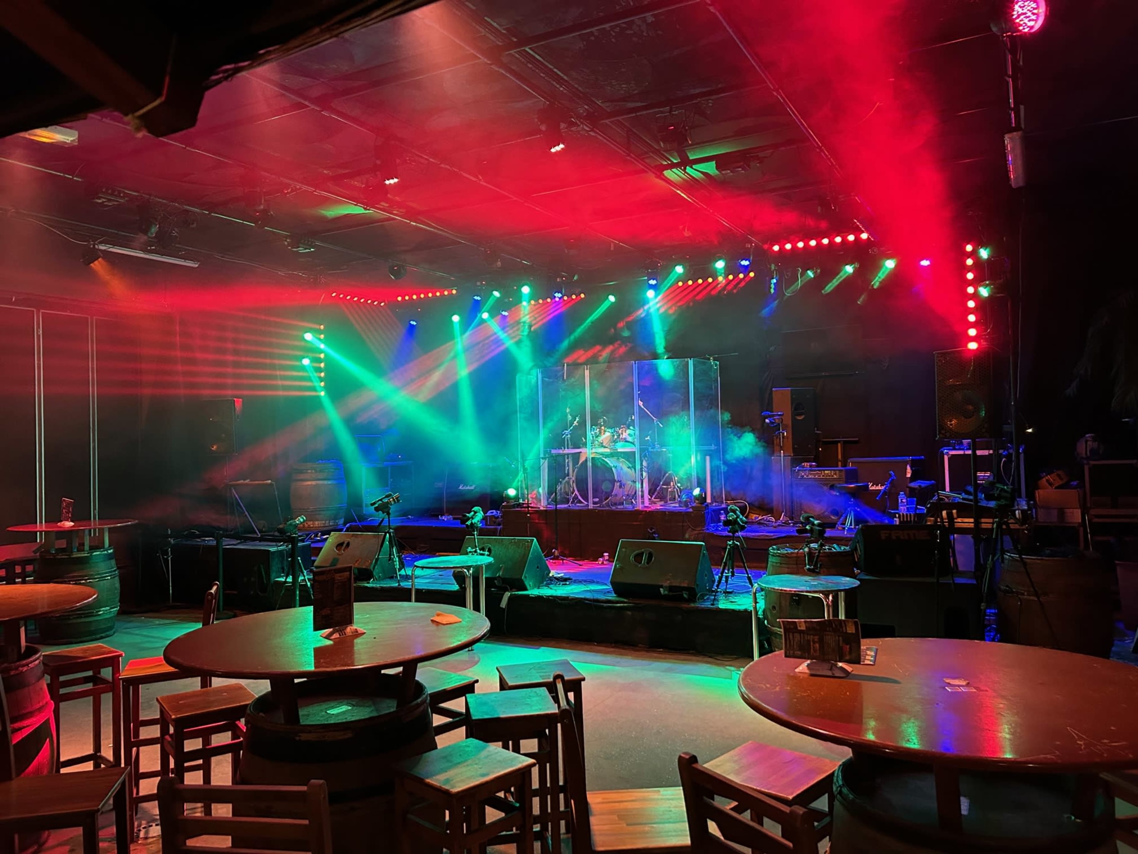 A stage setup with bright multicolored lights, fog effects, and musical instruments prepared for a performance, surrounded by empty tables and chairs.
