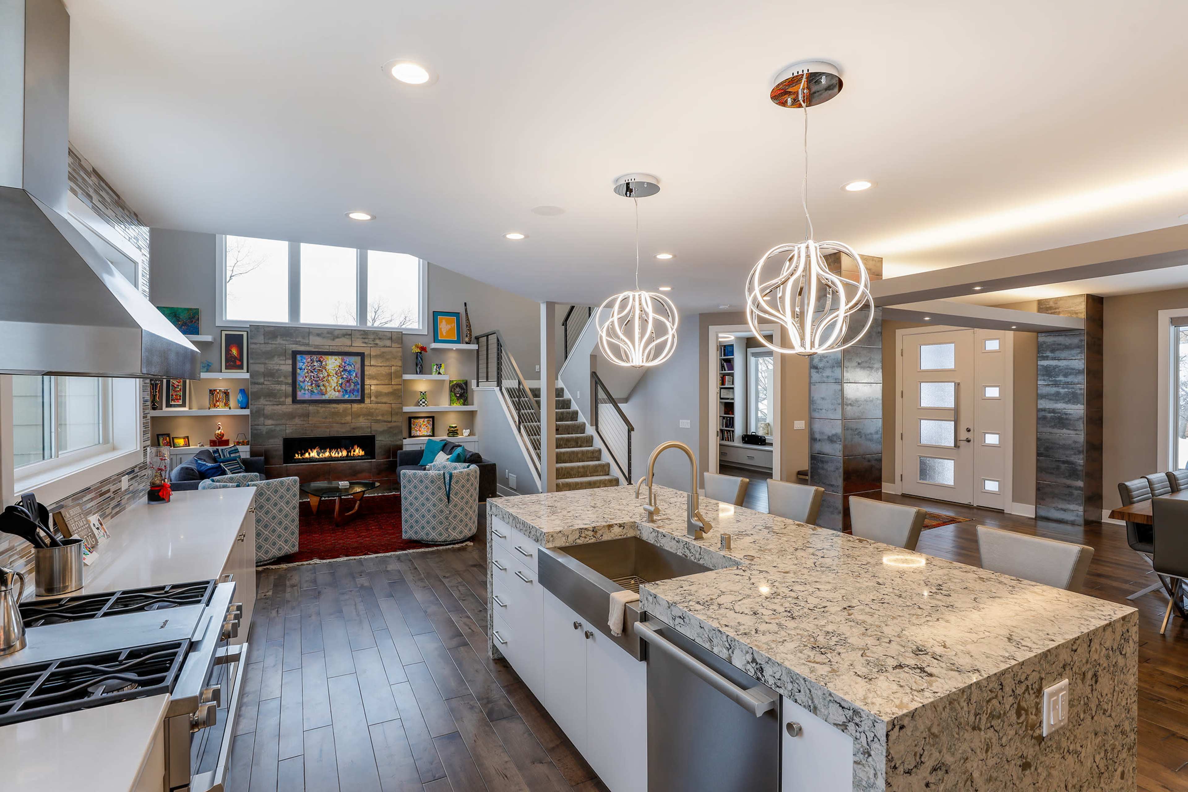 A modern kitchen with a large island, stainless steel appliances, and a living area featuring a fireplace and staircase.