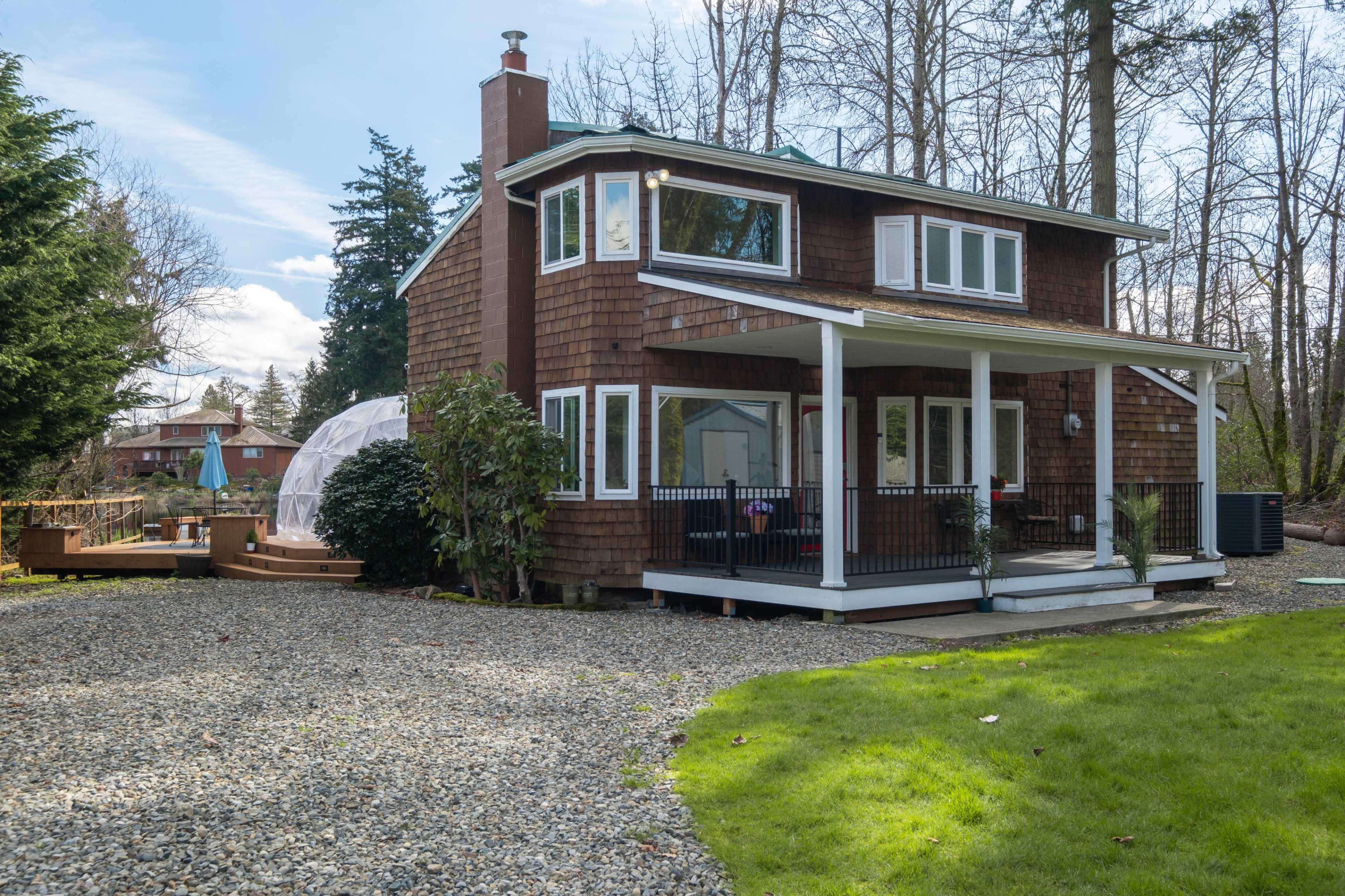 The image shows a two-story wooden house with a front porch, surrounded by gravel and a grassy lawn, set among trees.