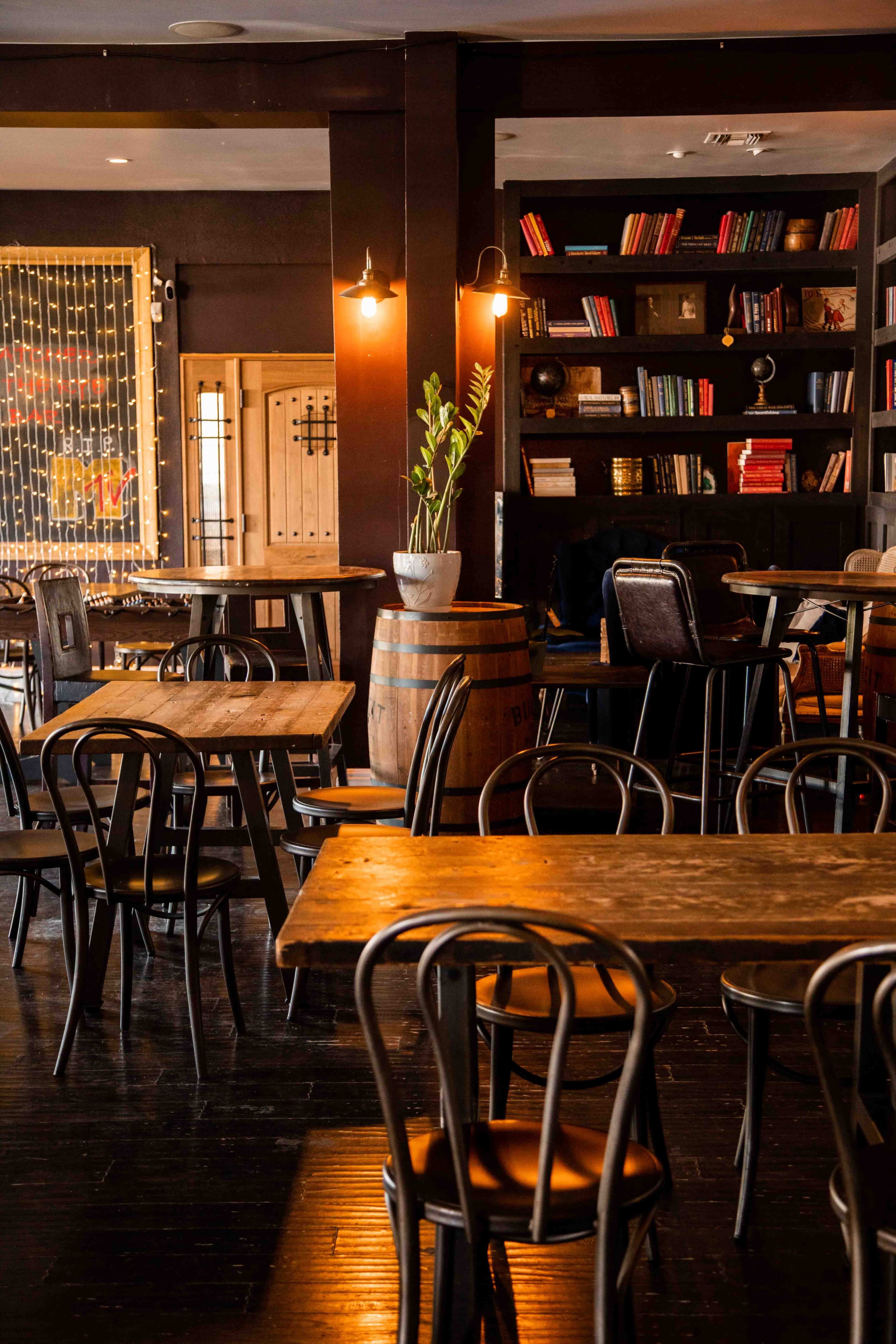 The image depicts a cozy café interior with wooden tables, black chairs, and shelves lined with books, featuring a barrel and a potted plant in the corner.