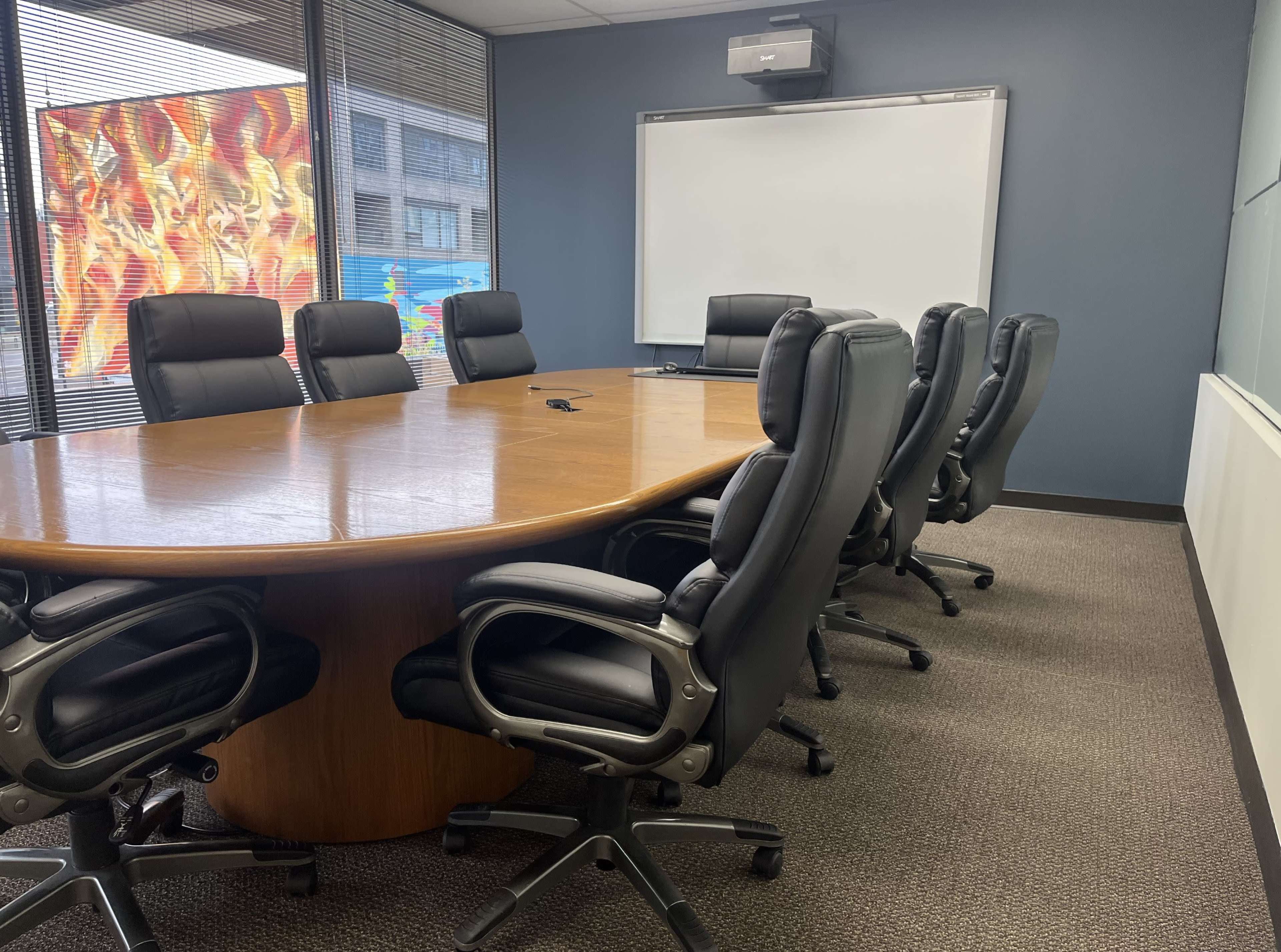 The image shows a conference room with a large wooden table surrounded by several black ergonomic chairs and a wall-mounted presentation screen.
