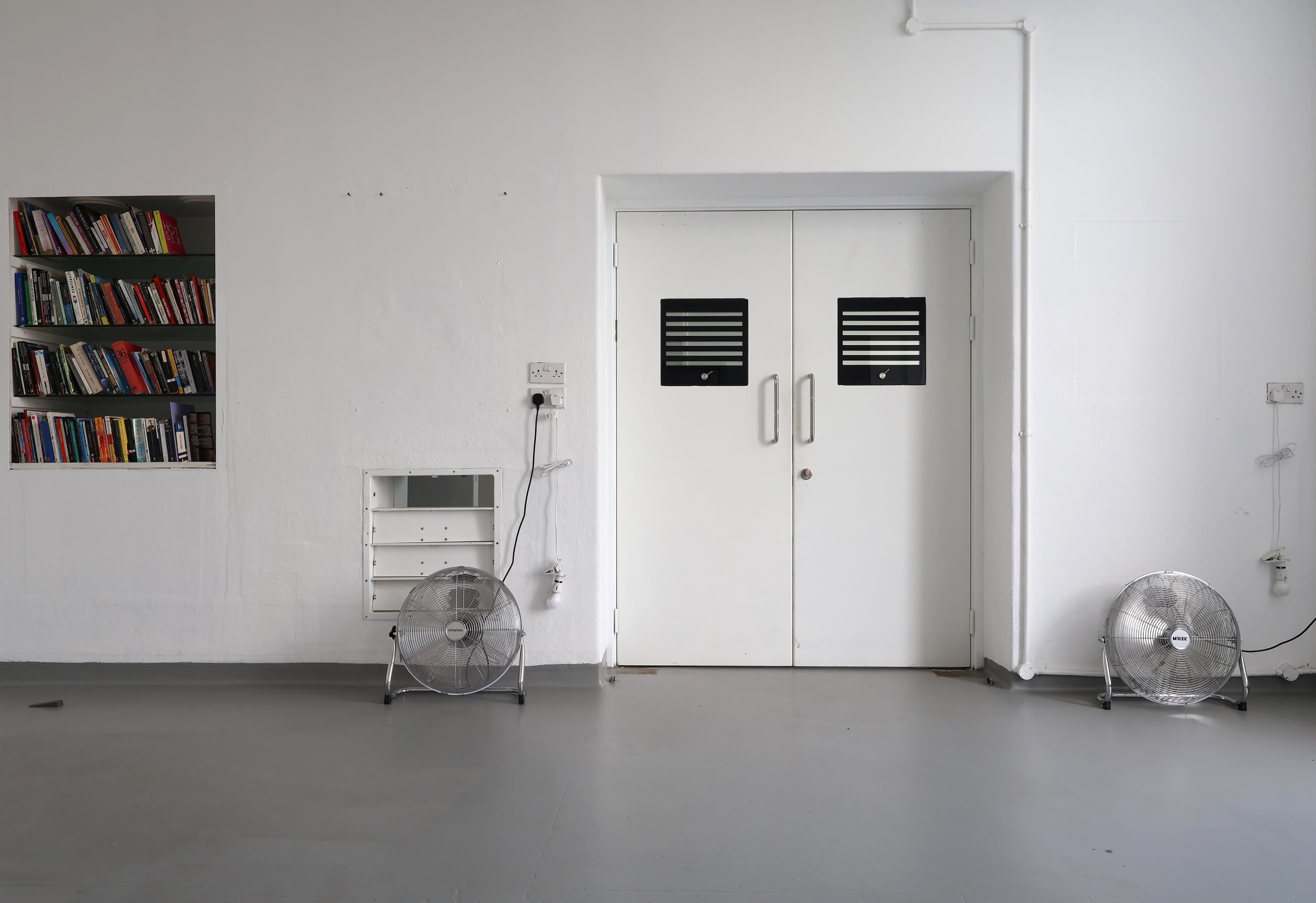 The image shows a minimalist room with a bookshelf on the left, two standing fans on the floor, and a set of double doors in the center.