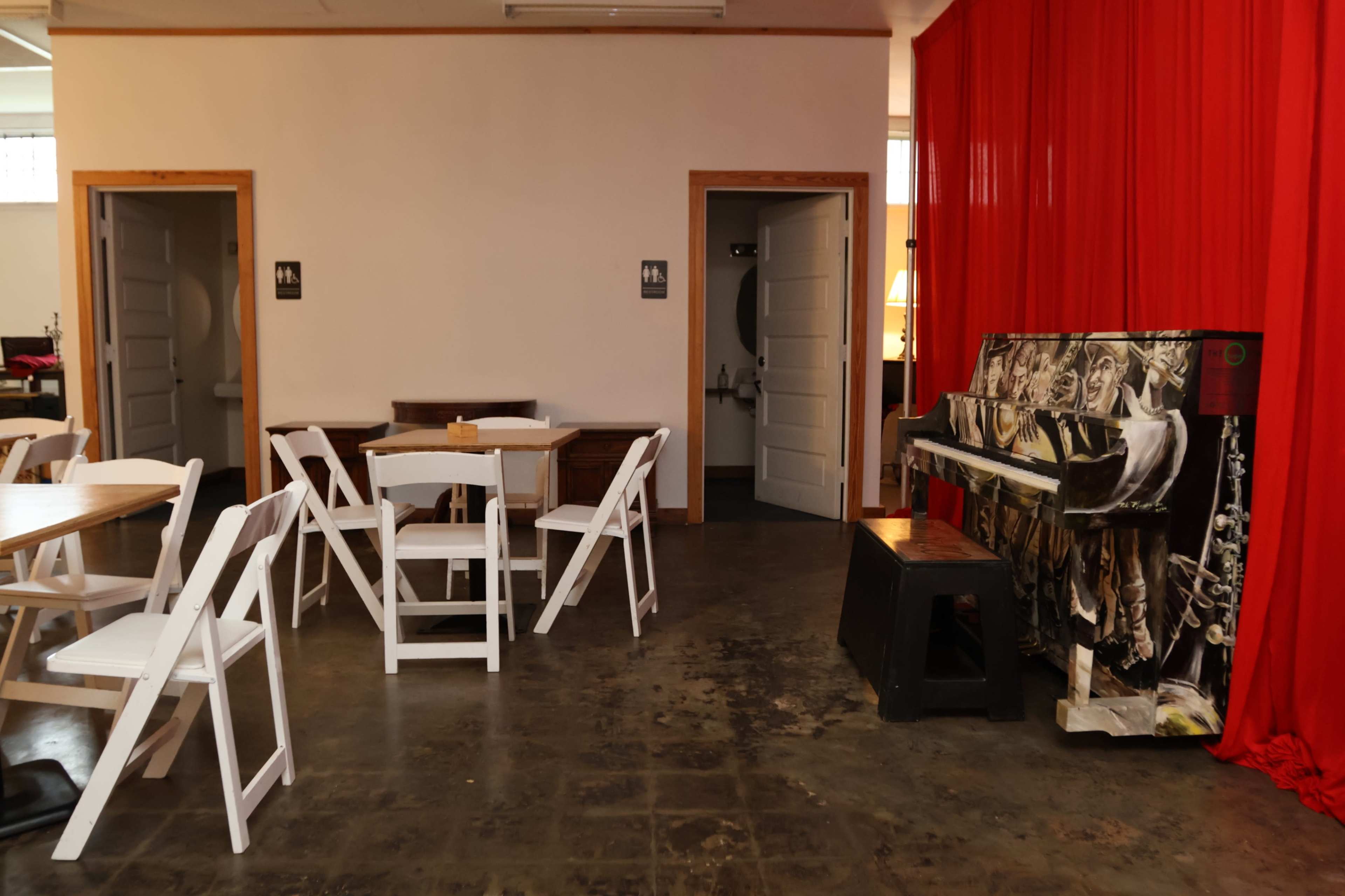 The image shows a sparse room with white folding chairs arranged around wooden tables, a piano decorated with art, and a red curtain hanging on one wall.