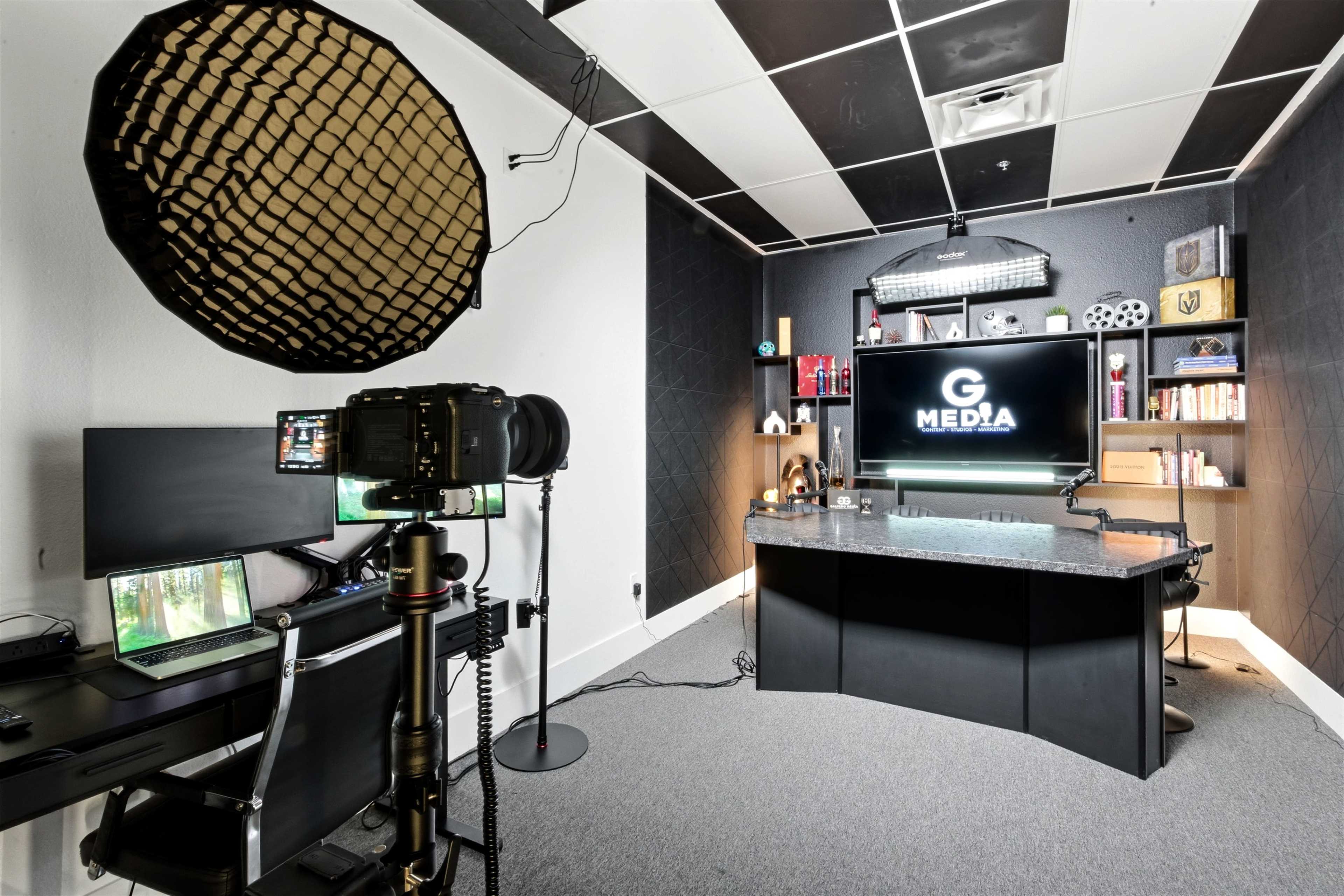 The image shows a modern media production studio featuring a black desk with a large screen displaying "G MEDIA," a camera on a tripod, and various equipment arranged on shelves.