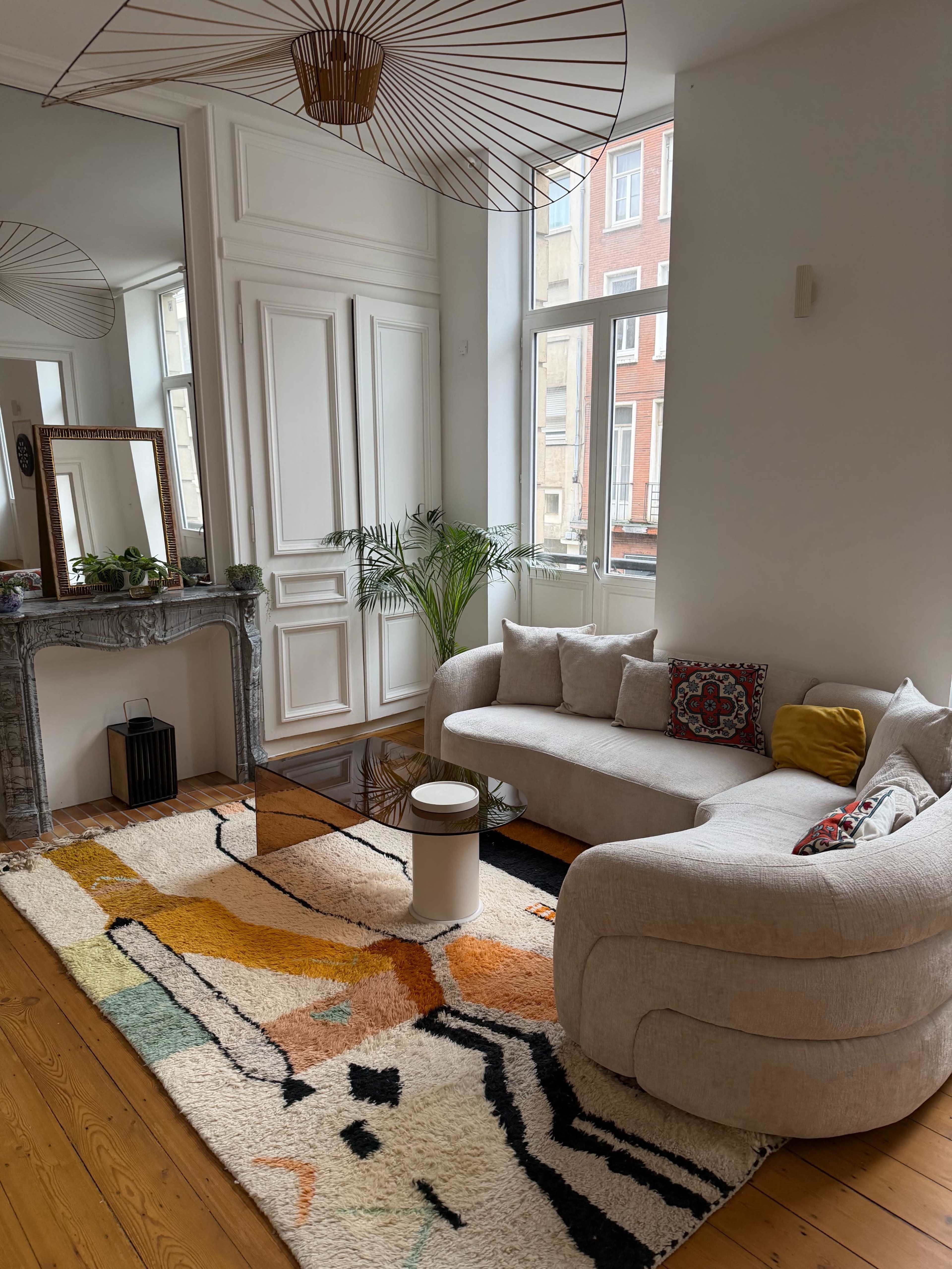 The image shows a cozy living room with a curved sofa, a patterned rug, and large windows allowing natural light to enter.