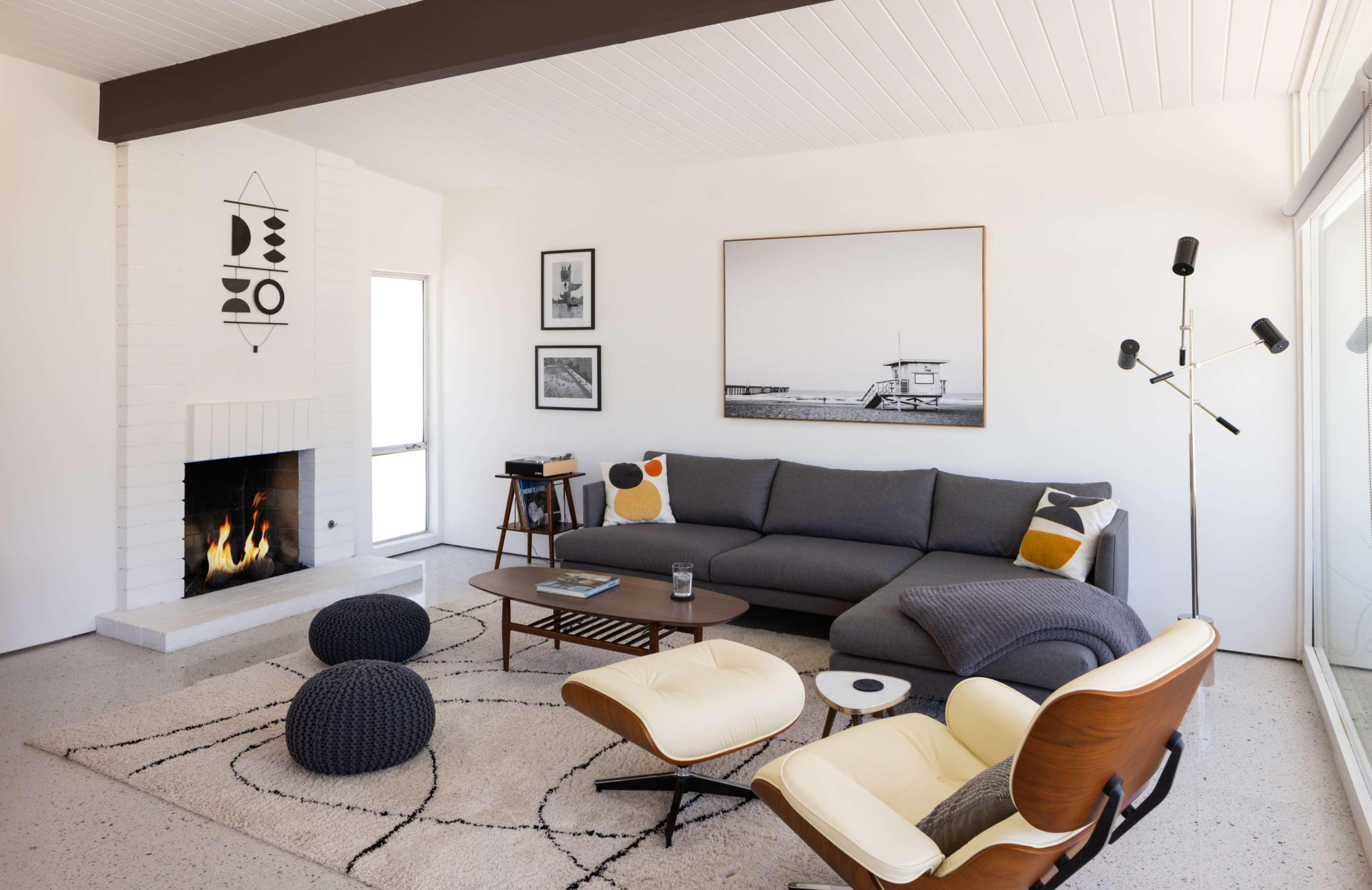 The image shows a modern living room with a gray sectional sofa, a round coffee table, and two upholstered ottomans, featuring a fireplace and large windows that provide natural light.