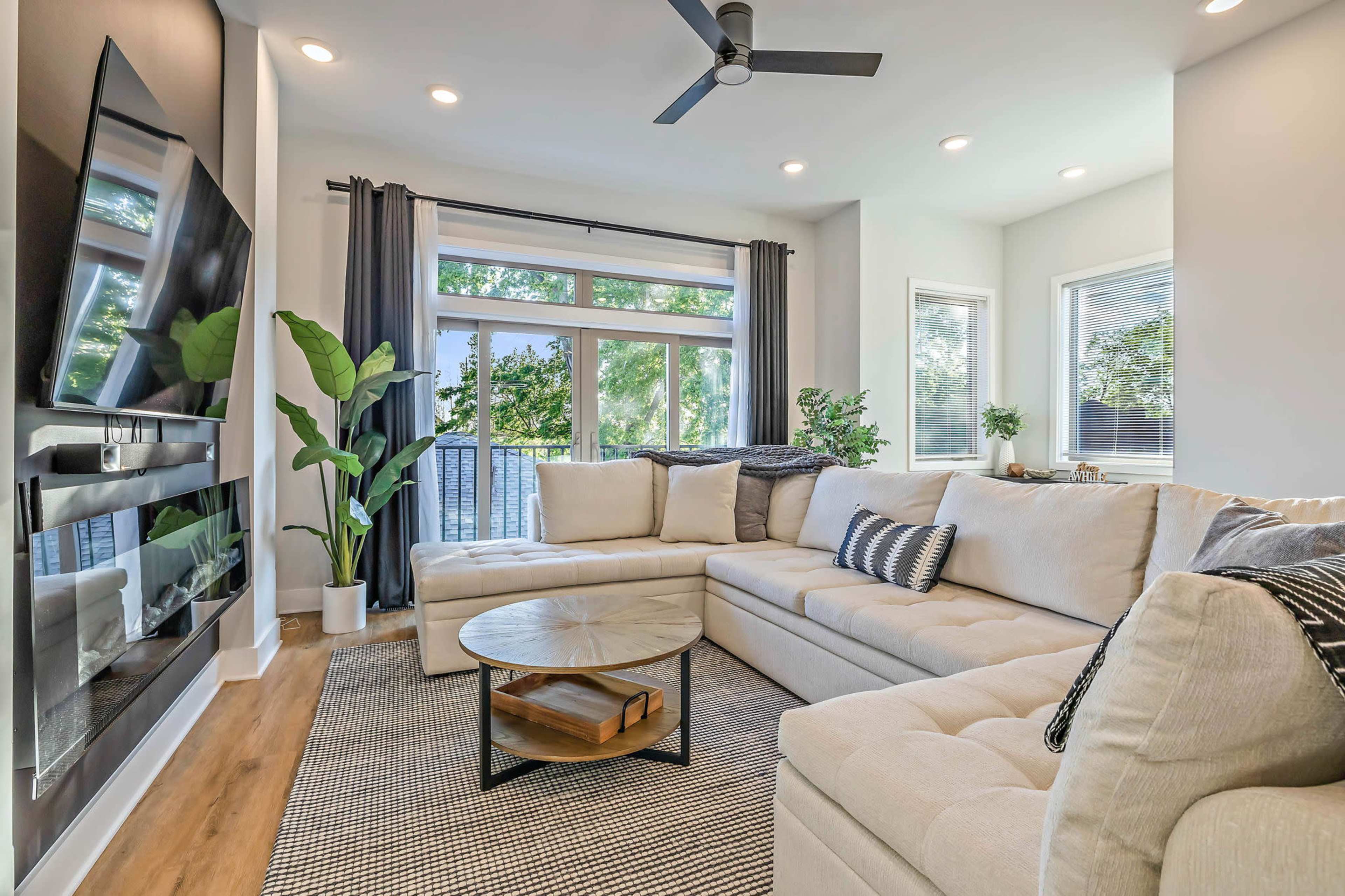A modern living room features a large sectional sofa, a round coffee table, and a wall-mounted television, complemented by natural light streaming through large windows.