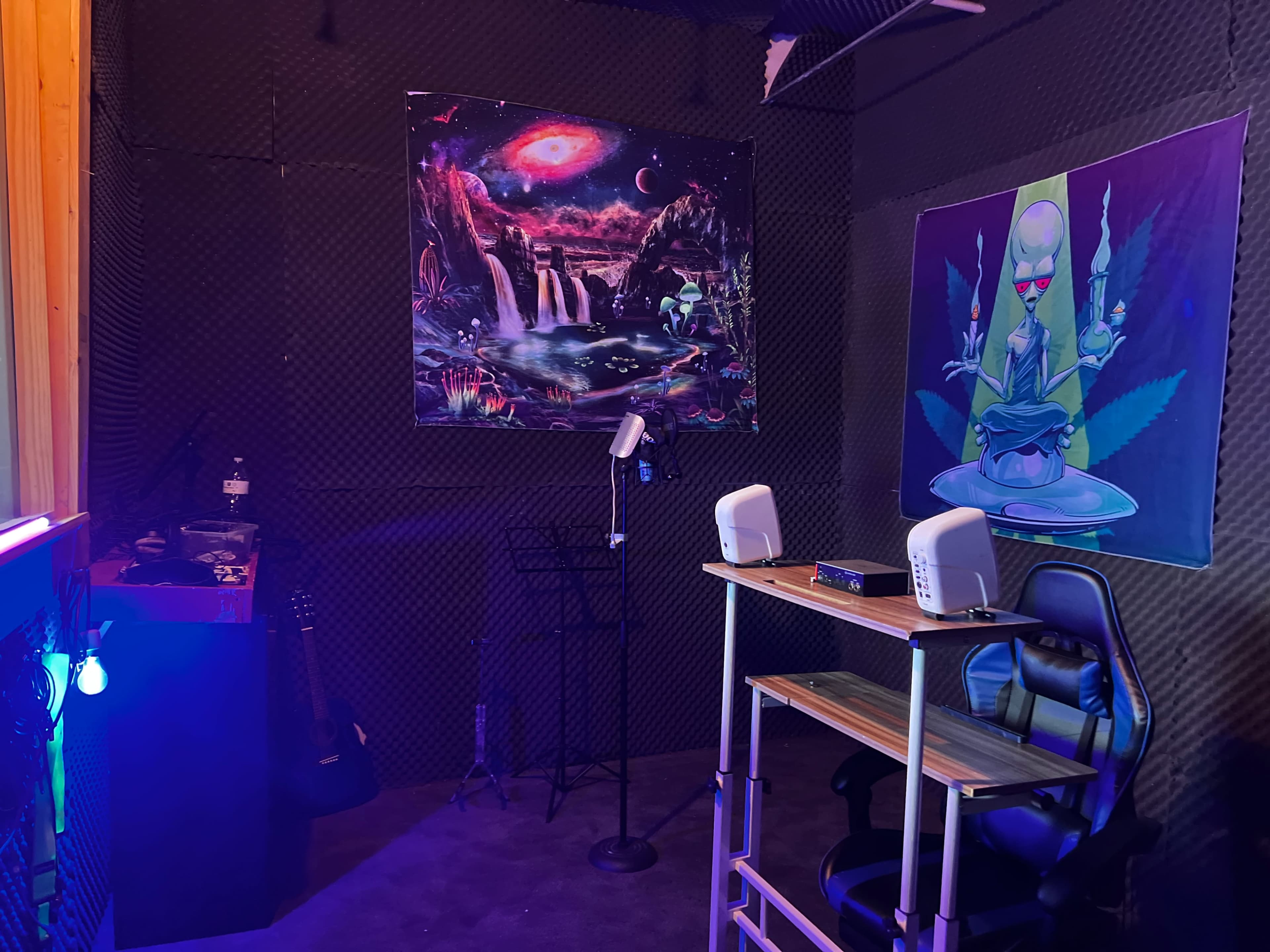 The image depicts a studio space featuring acoustic foam walls, a desk with a chair, a microphone stand, and two colorful posters on the walls.