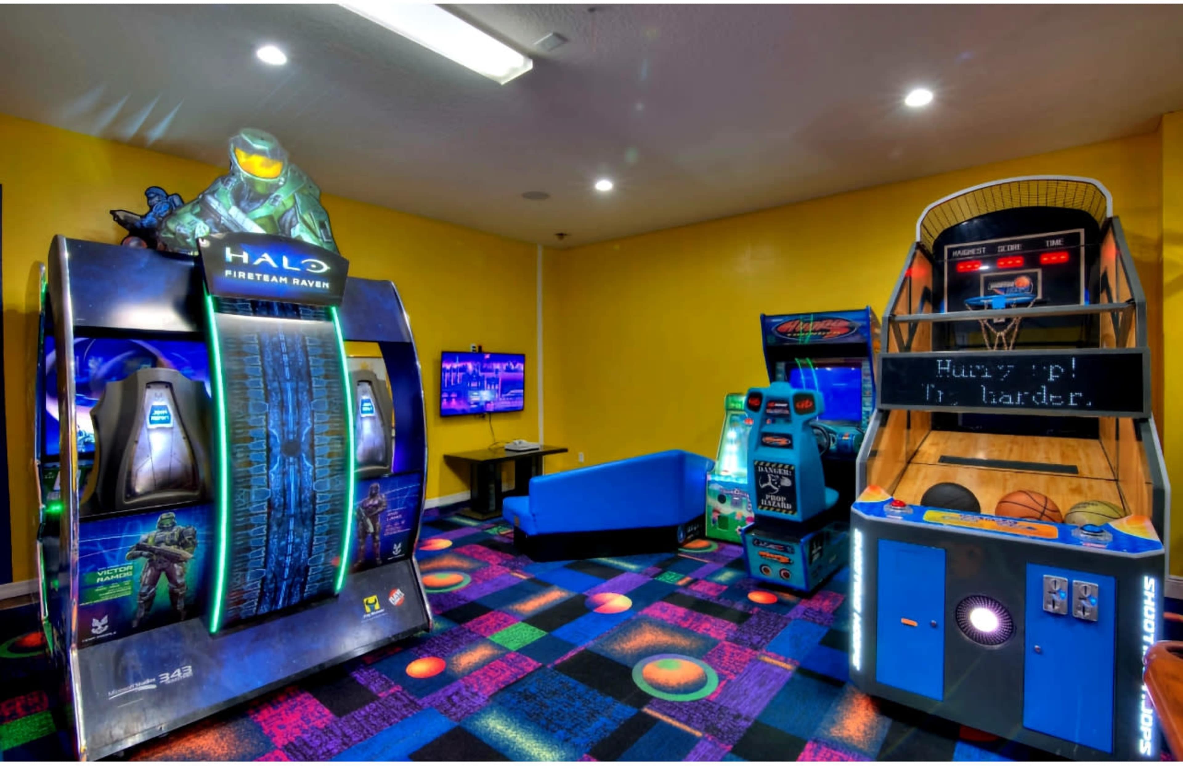 A brightly colored arcade room featuring a "Halo" video game machine, a basketball arcade game, and a television.