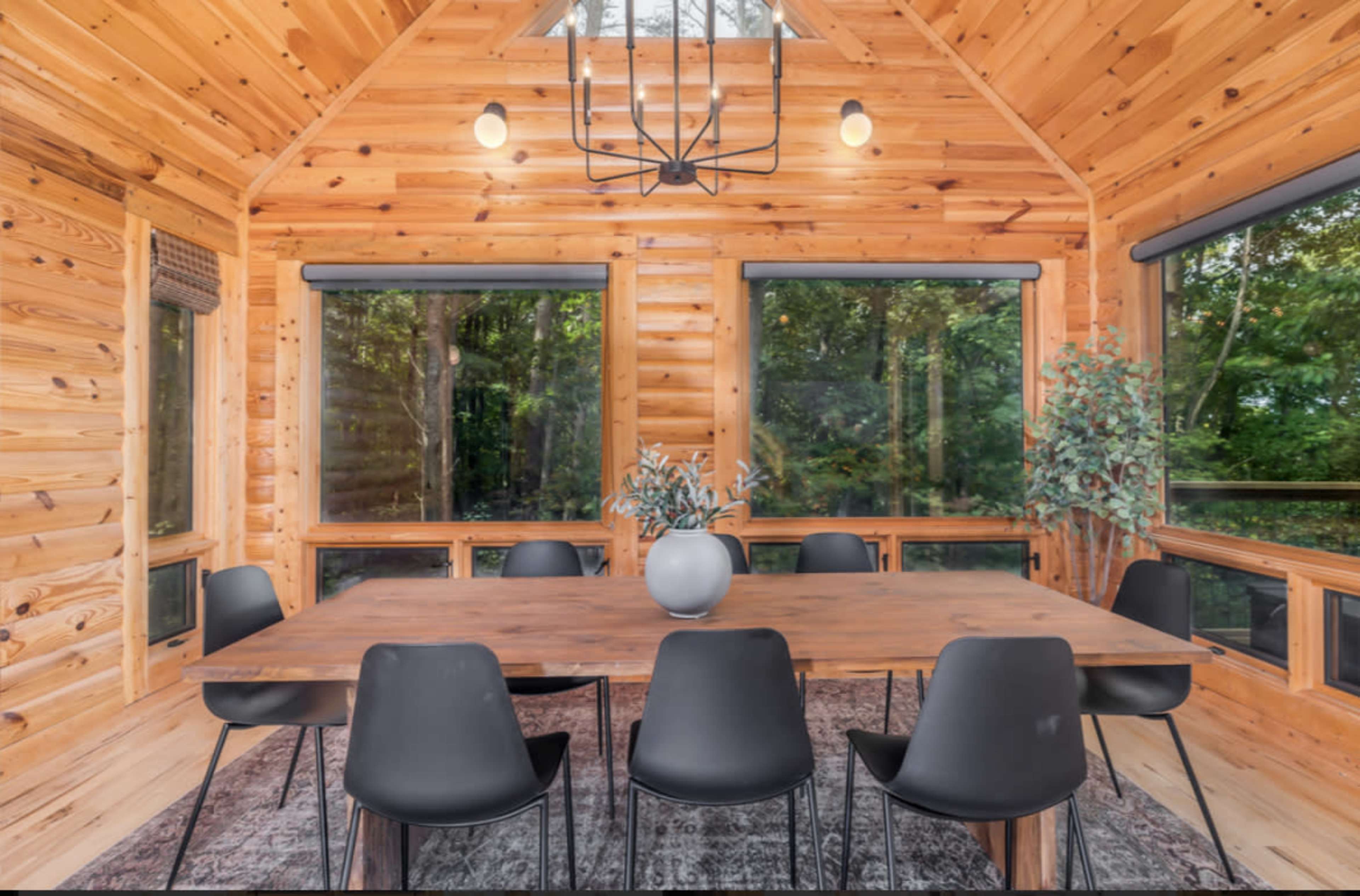 A wooden dining area features a large table surrounded by black chairs, with ample natural light coming through large windows.