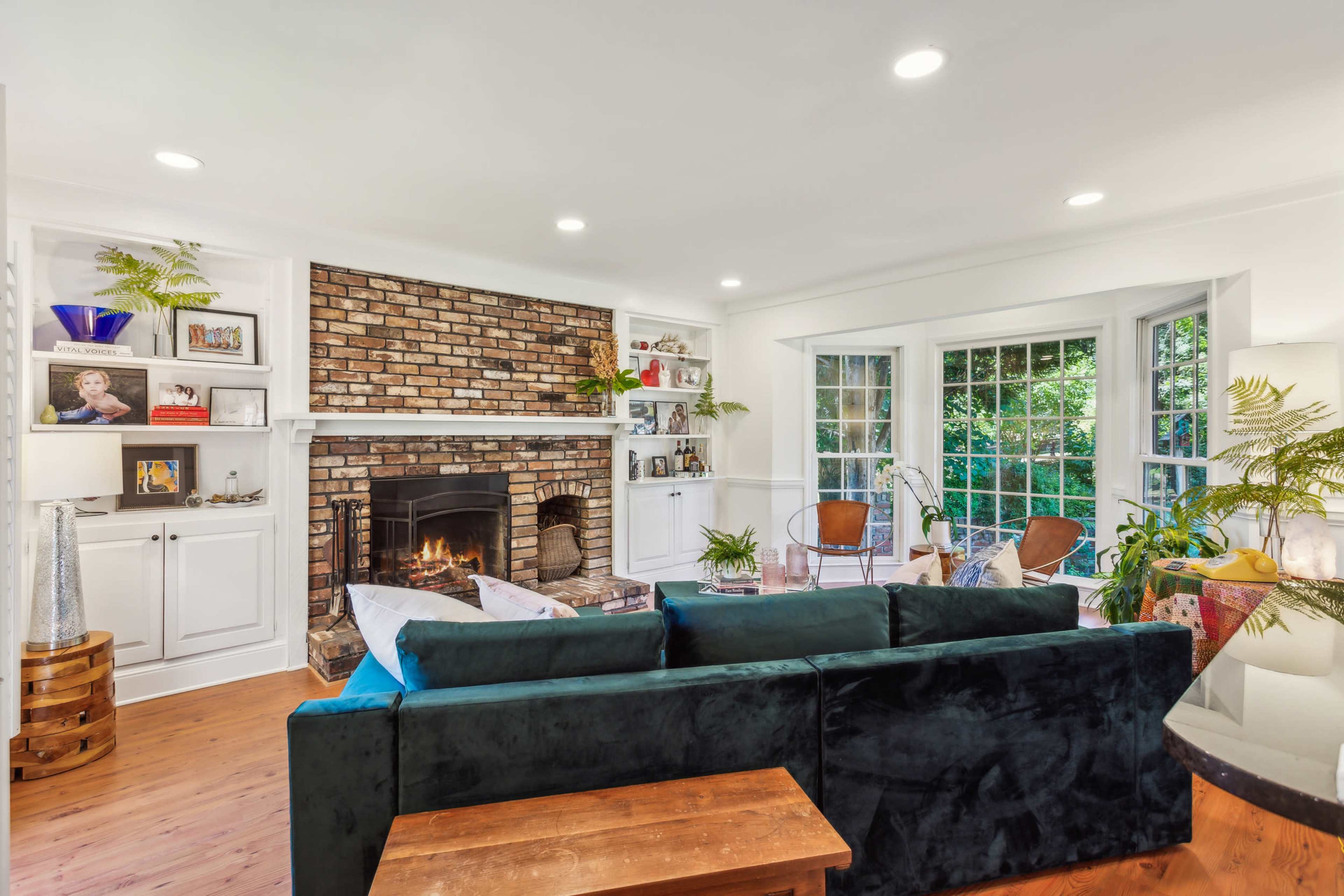 The living room features a brick fireplace with built-in shelves, a dark sofa, and large windows that overlook a garden.
