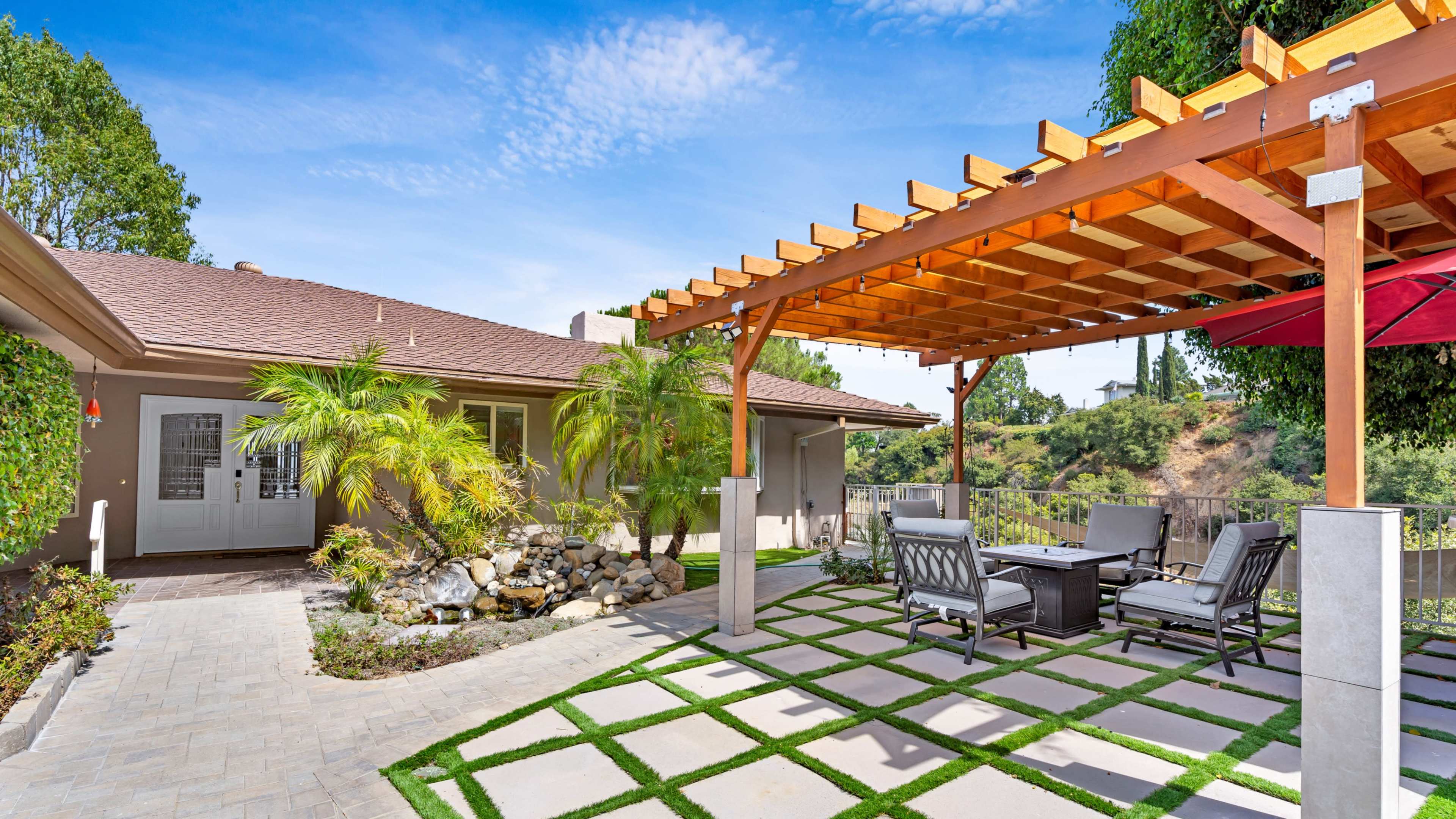 A covered patio with outdoor furniture is situated beside a house, surrounded by greenery and stone landscaping.