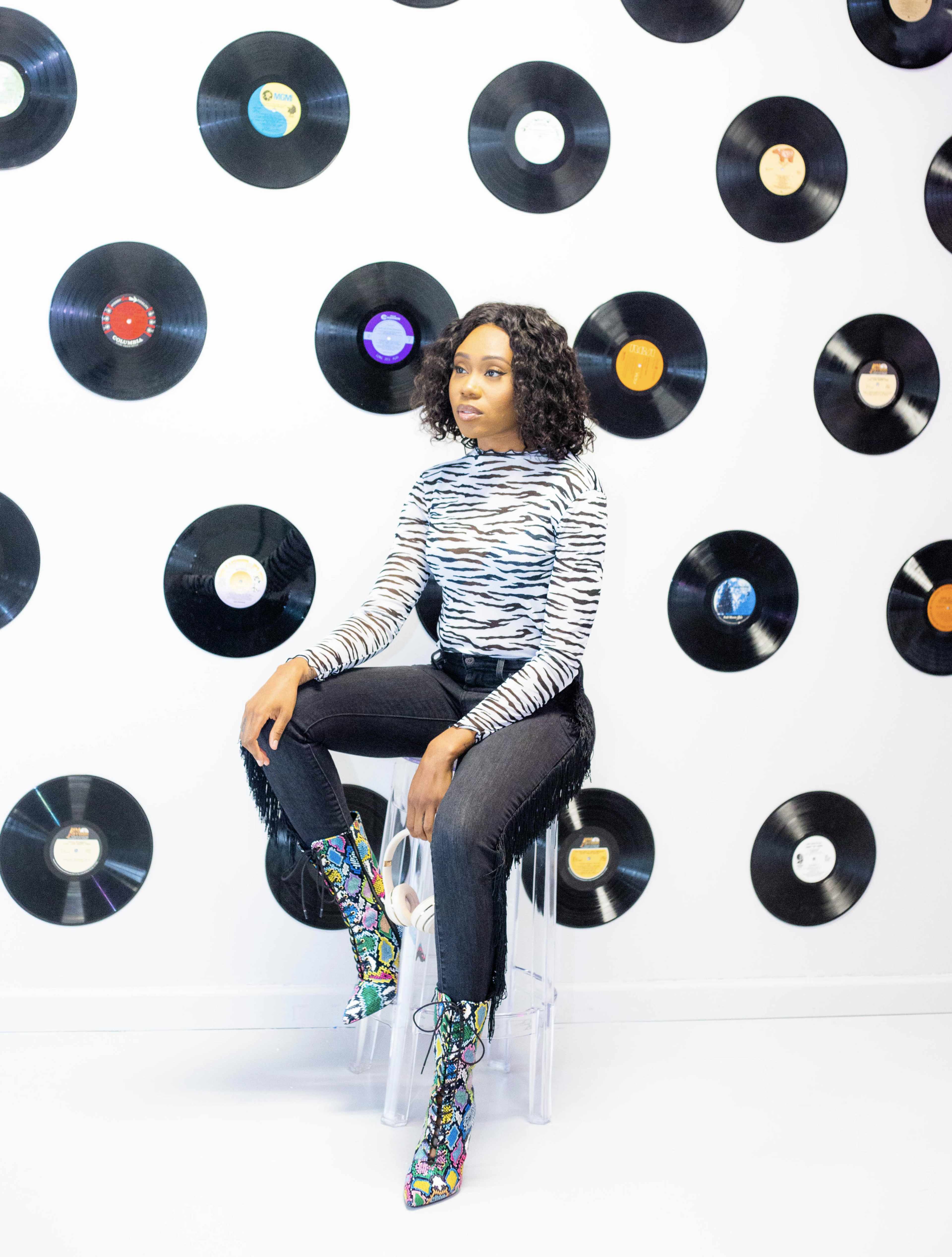 A person sits on a clear stool in front of a wall adorned with black vinyl records while wearing a striped top and colorful boots.