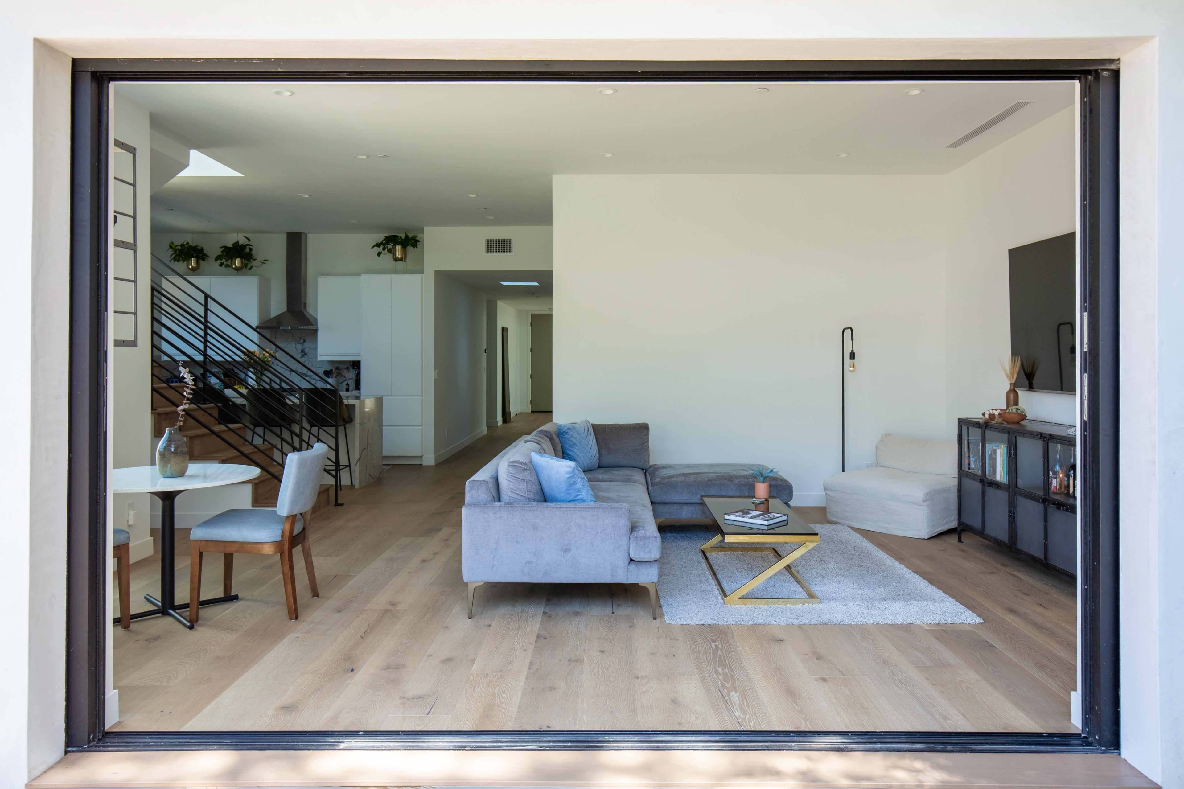 A large sliding glass door reveals a modern living space with a sectional sofa, a coffee table on a rug, and a dining area with a small table.