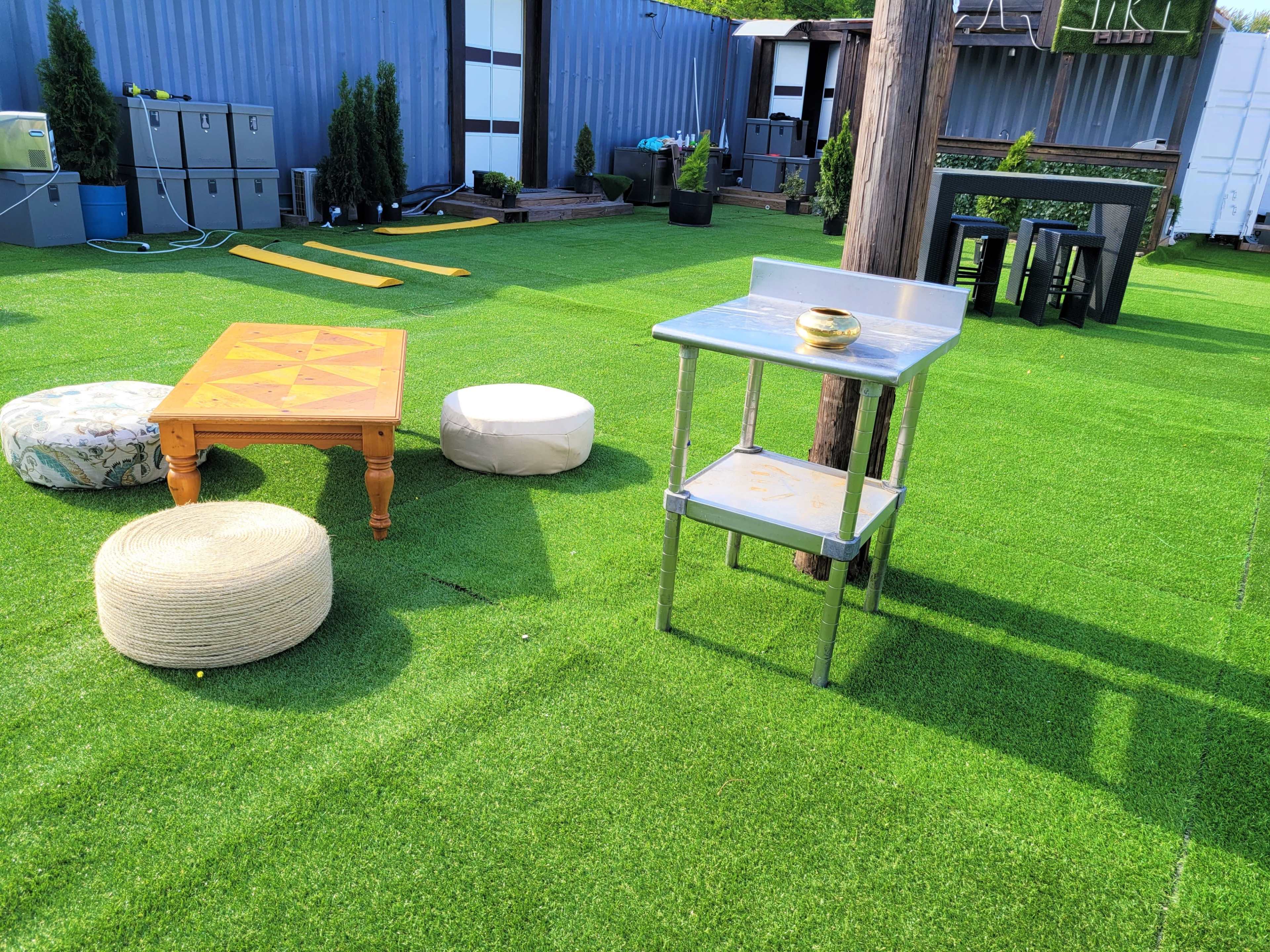 The scene features a green artificial grass area with a wooden table, round cushions, and a metal kitchen prep table.