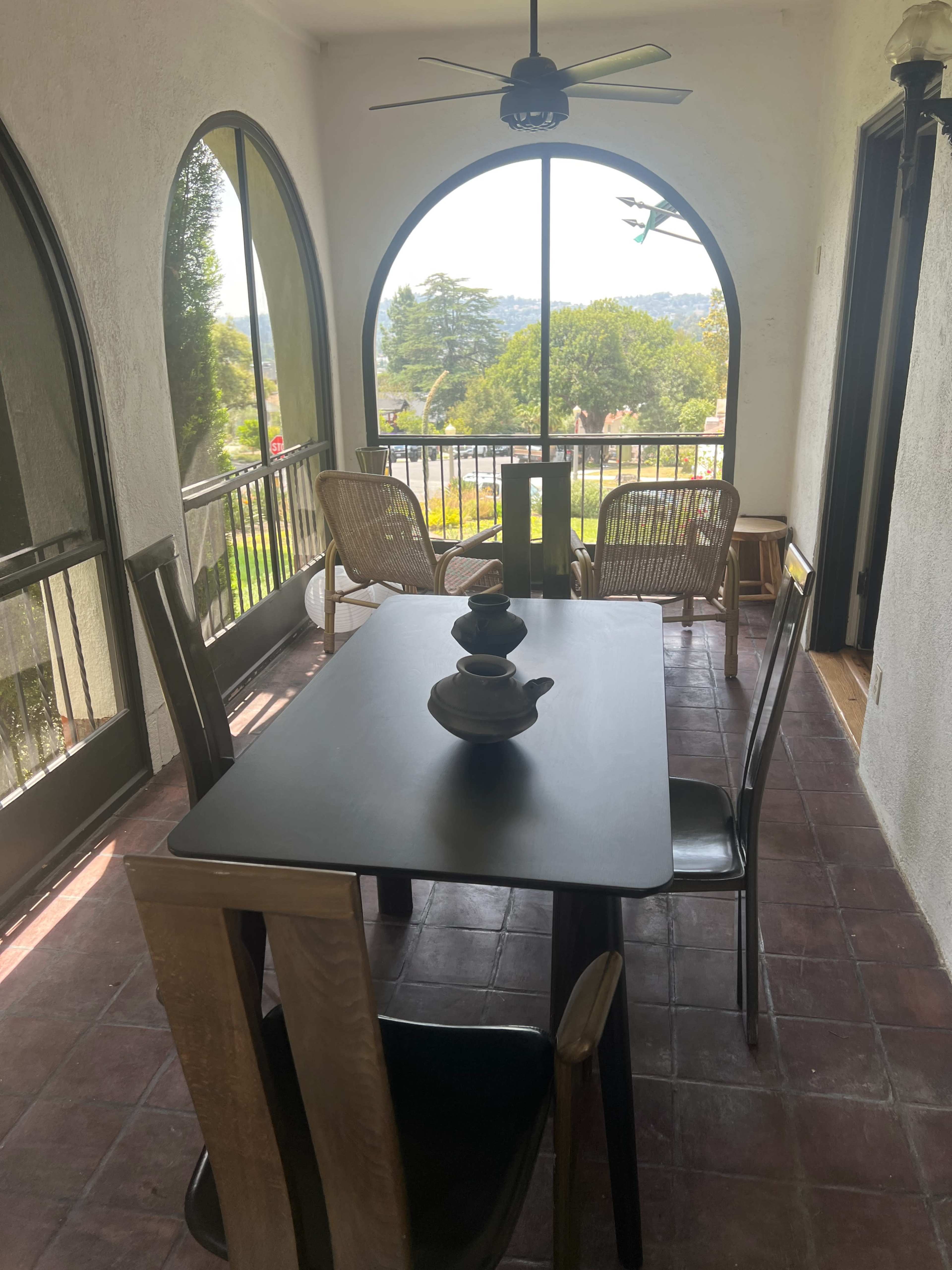 The image shows a tiled patio with a black dining table, four chairs, a teapot on the table, and large arched windows revealing a view of greenery outside.