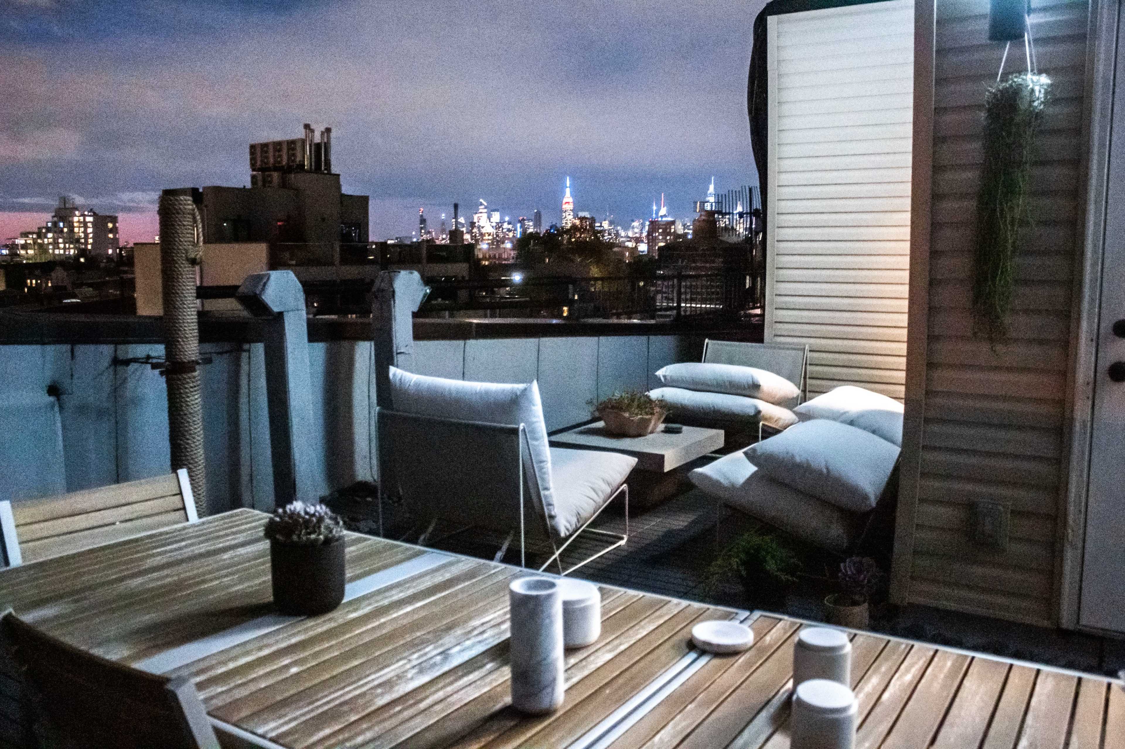 A rooftop terrace features a wooden table with candles and minimal seating, overlooking a skyline with illuminated buildings at night.