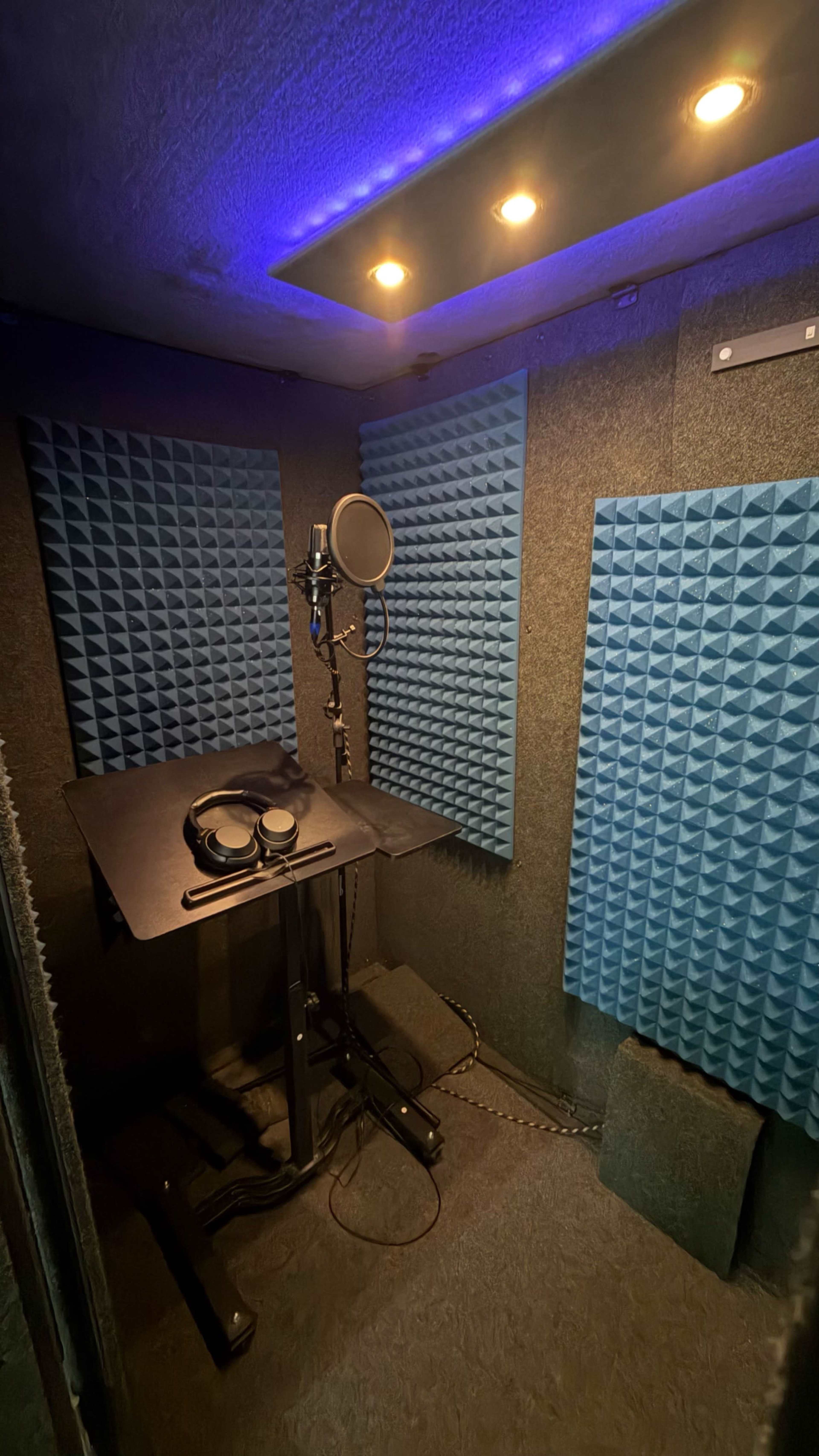 The image shows a vocal recording booth equipped with soundproof blue foam panels, a microphone on a stand, headphones, and a small black table.