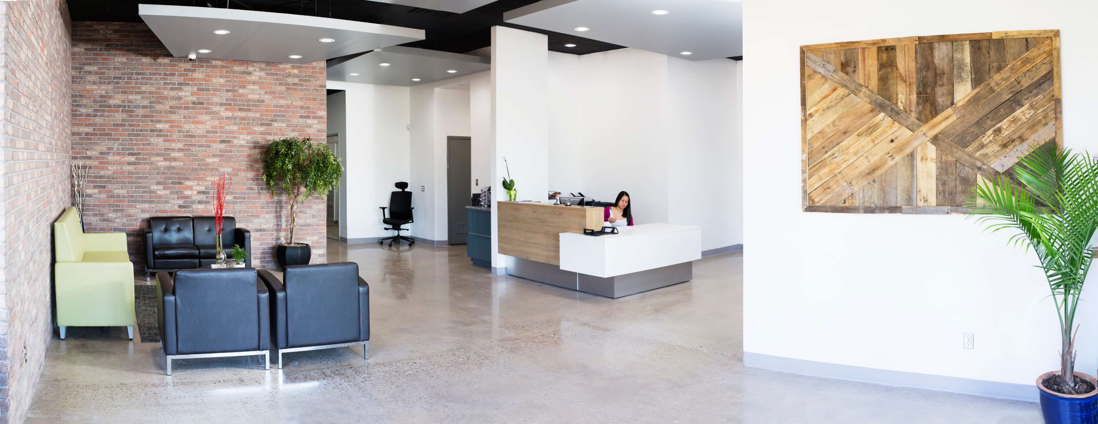 A modern reception area featuring a combination of brick and white walls, with seating arrangements that include green and black sofas, a wooden reception desk, and a potted plant in the corner.