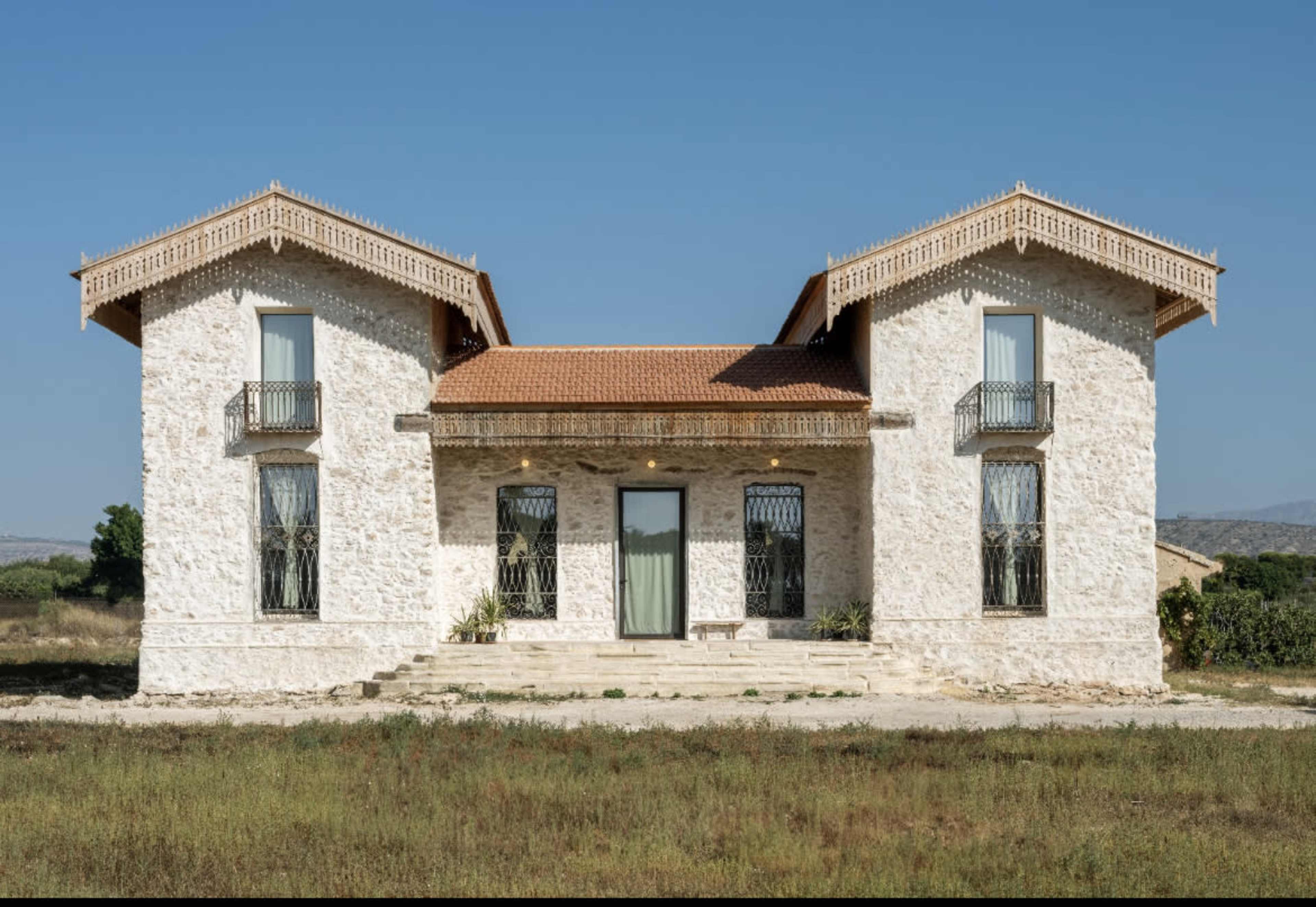 A symmetrical, two-story stone house with balconies and a red-tiled roof sits in a grassy field under a clear blue sky.